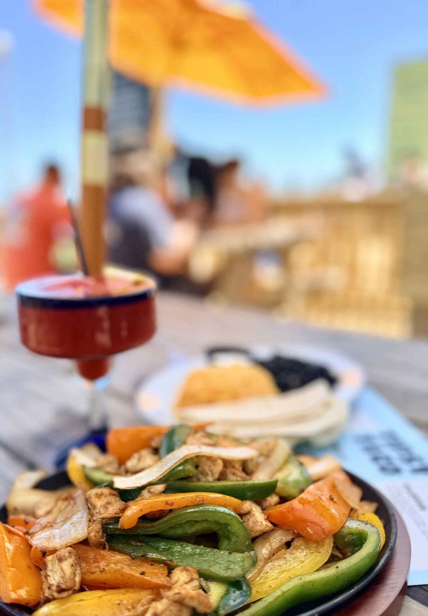 Close-up of a plate with stir-fried vegetables and chicken, including green, yellow, and orange bell peppers, broccoli, and chicken strips, in the foreground. In the blurred background, there are people sitting at a table outdoors, with a red soy sauce dispenser and a bowl of dipping sauce, with a blue sky visible.