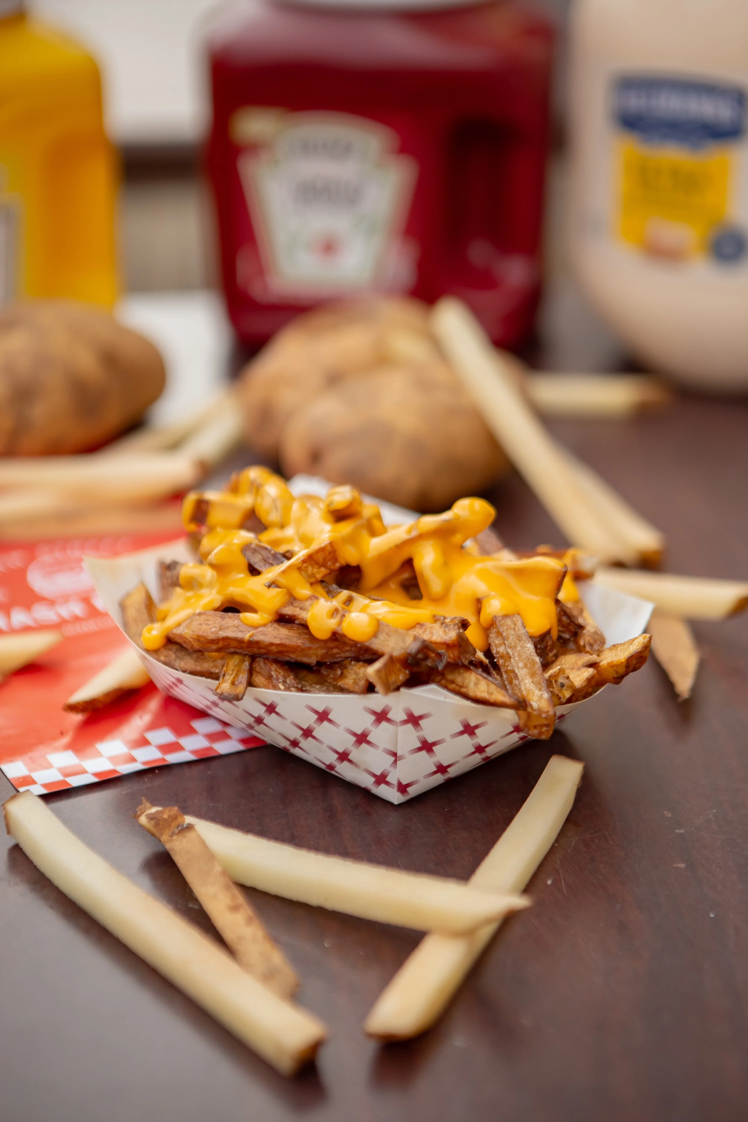 A serving of loaded fries topped with cheese, chopped beef, and bacon bits. In the background, there are baked potatoes, packets of mustard, ketchup, and mayonnaise on a table.