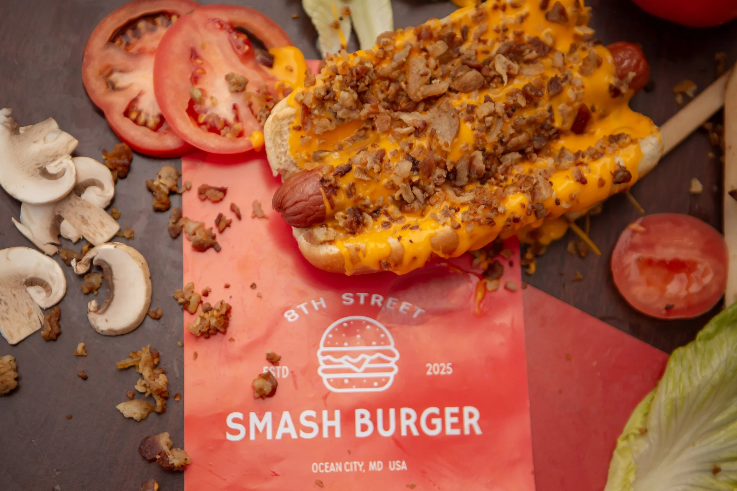Close-up of a Smash Burger with cheese and ground beef, surrounded by sliced tomatoes, mushrooms, lettuce, and other vegetables on a dark surface.