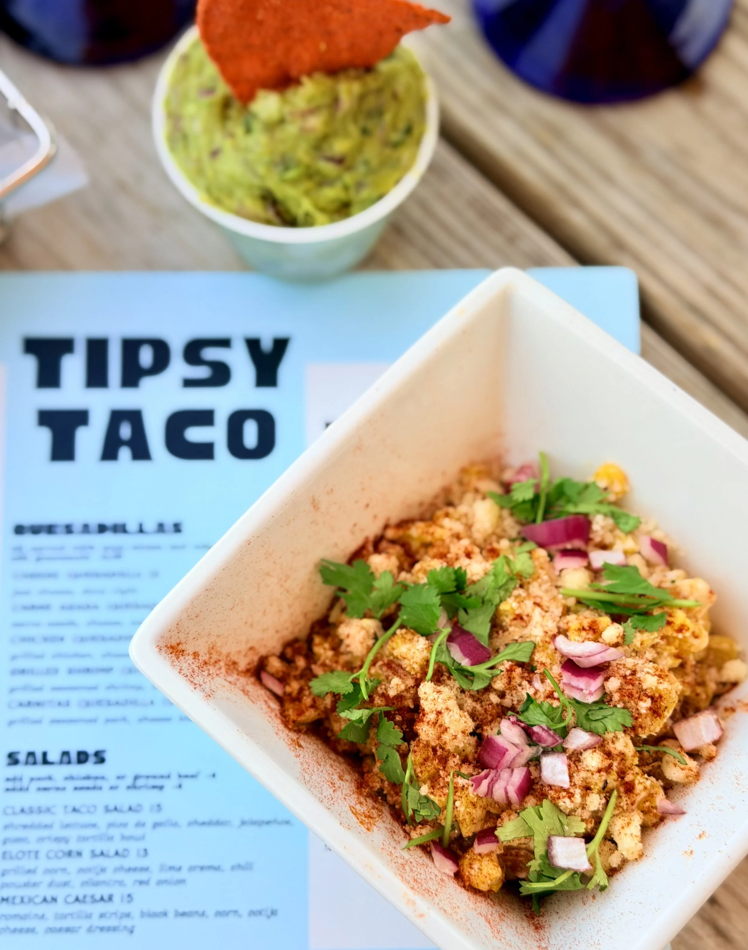 A white dish with diced ingredients topped with cilantro, red onion, and garnished with lime and a sprinkle of paprika, placed on a blue and white menu that includes text about tacos and salads.