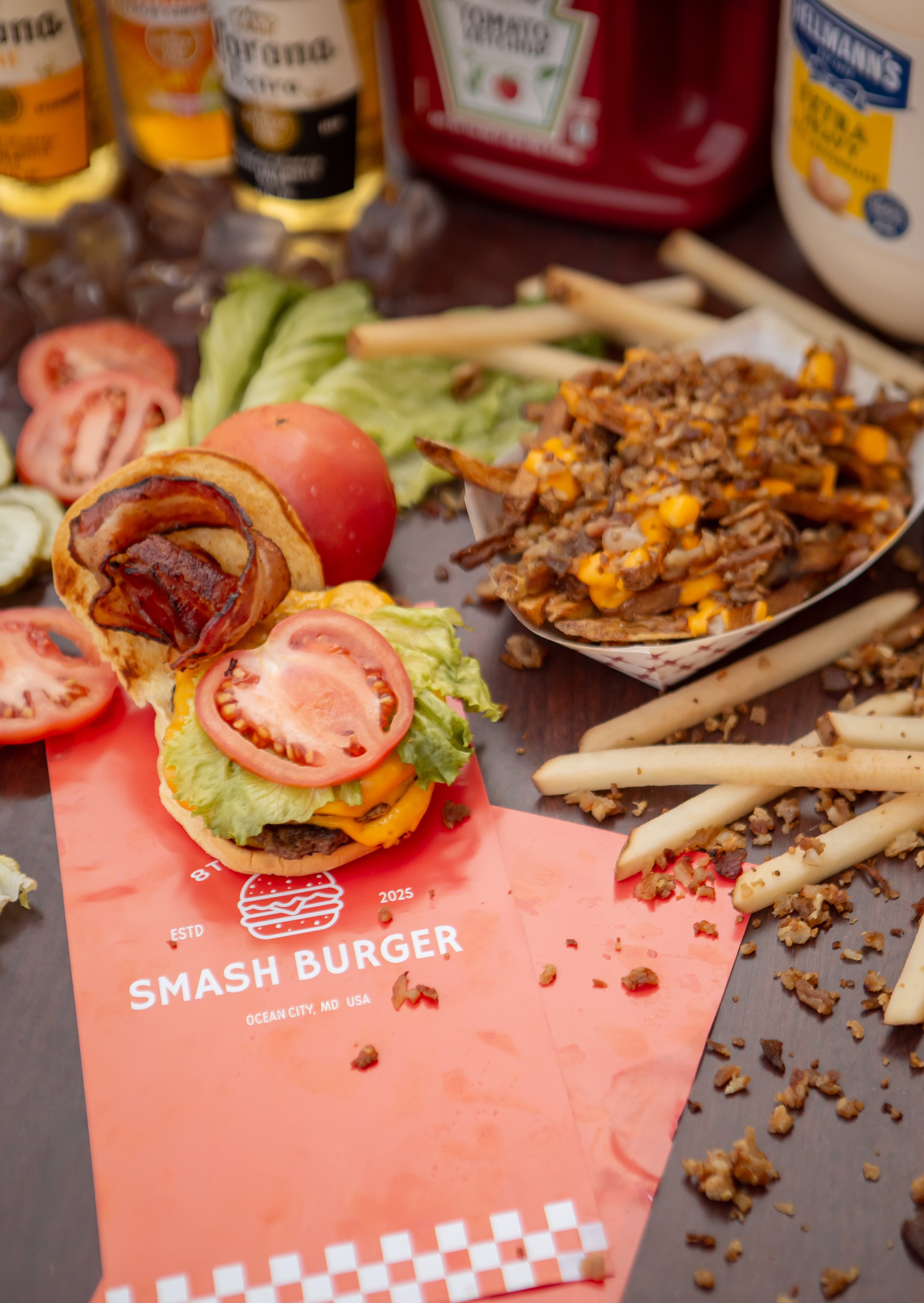 Close-up of a tray with a burger, tomato slices, chopped lettuce, and a side of seasoned fries with chopped meat, along with condiment bottles and packs in the background.