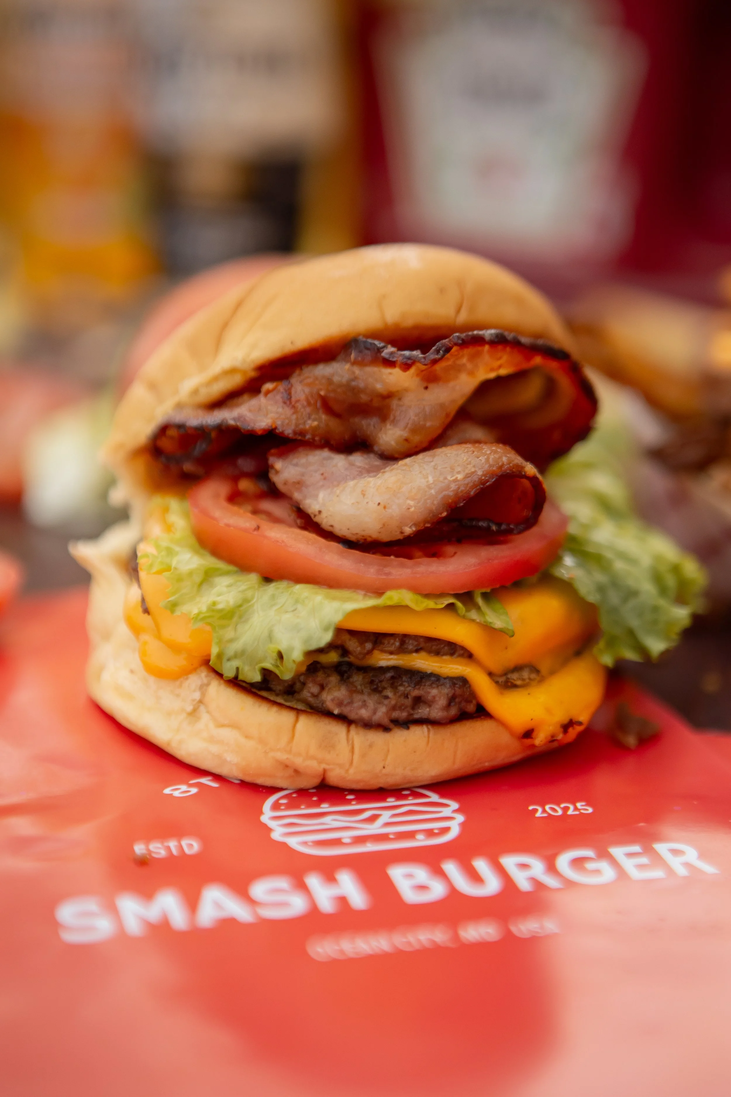 Close-up of a gourmet cheeseburger with lettuce, tomato, cheddar cheese, bacon, and a beef patty on a bun, with condiment bottles in the blurred background.
