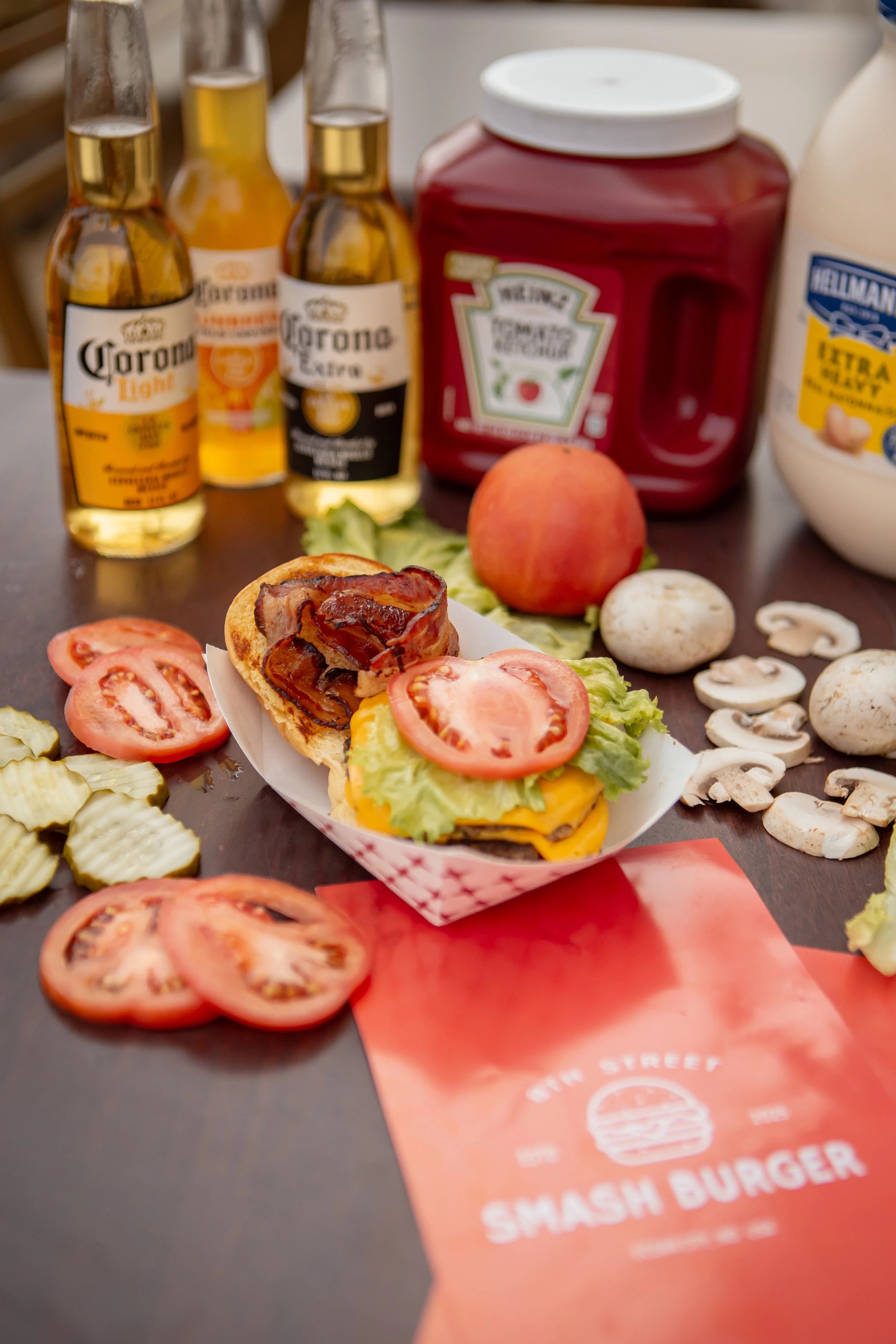 A sandwich with bacon, cheese, lettuce, and tomato slices in a paper tray, surrounded by sliced tomato and mushrooms on a table. In the background, there are three bottles of Corona beer, a ketchup bottle, and a mayonnaise bottle.