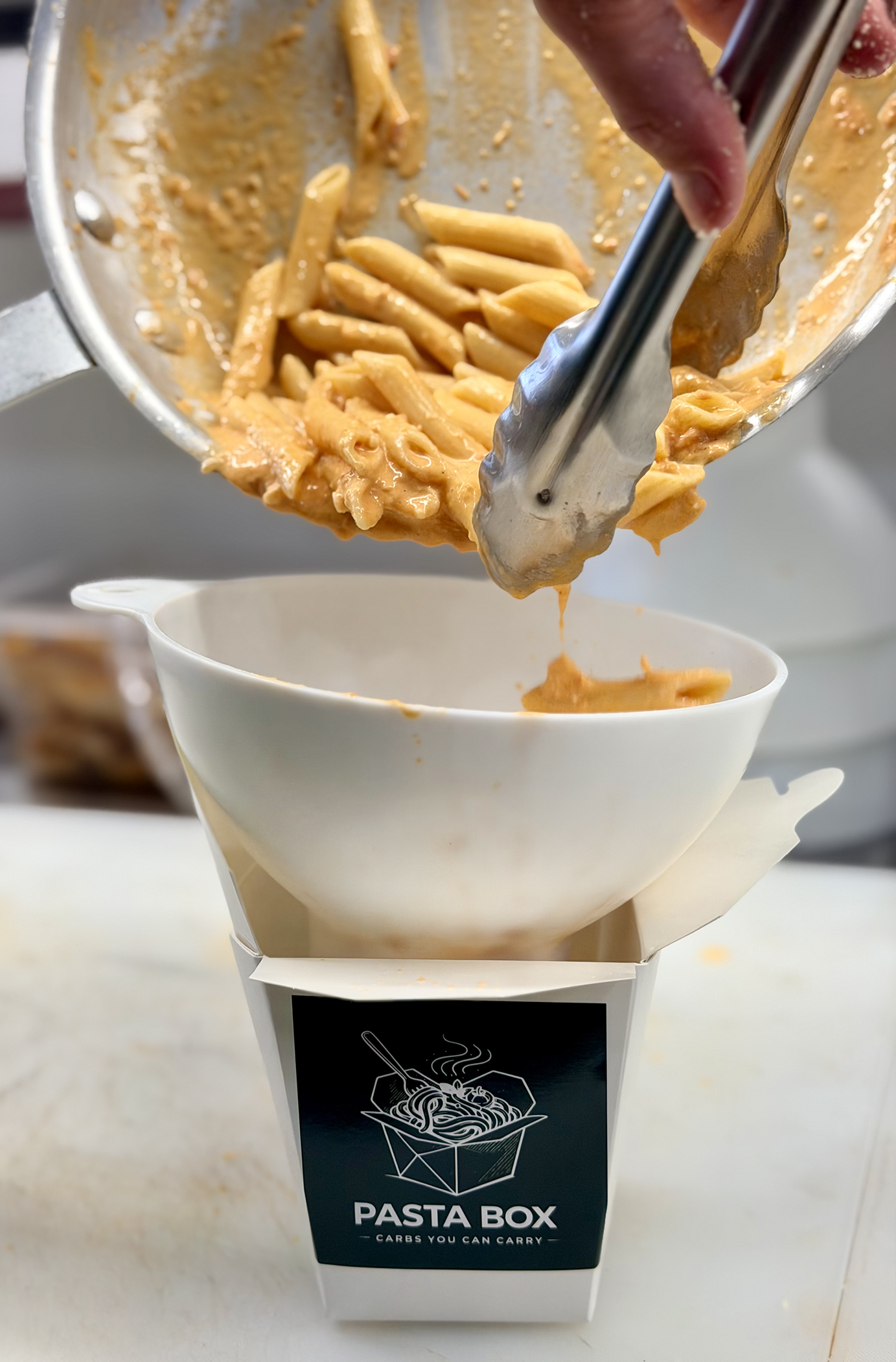 A white food container labeled "Pasta Box" with a graphic of a takeout box filled with pasta, placed on a white surface. The container is releasing cooked pasta from a silver cup into a white bowl.
