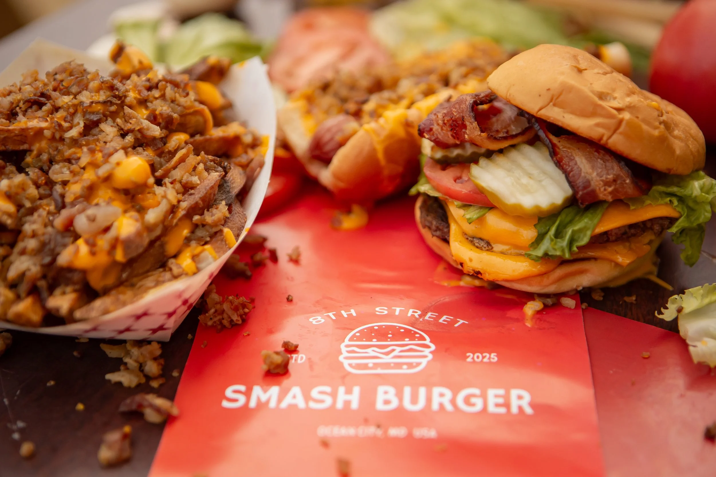 Close-up of a cheeseburger with lettuce, tomato, bacon, cheese, and a bun, next to a serving of chili or shredded meat with corn, on a red paper with a burger logo.