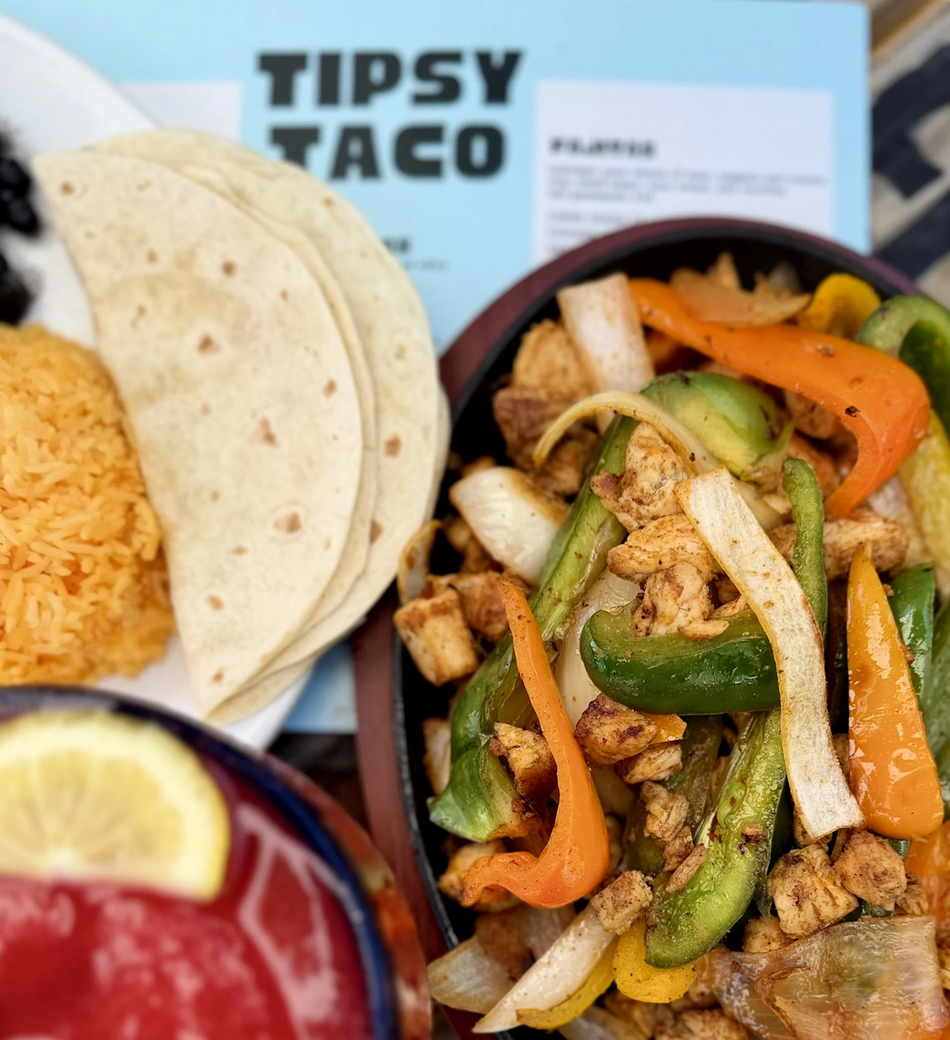 Close-up of a plate with various tacos, including chicken, vegetables, and cheese, with a Tipsy Taco box in the background.