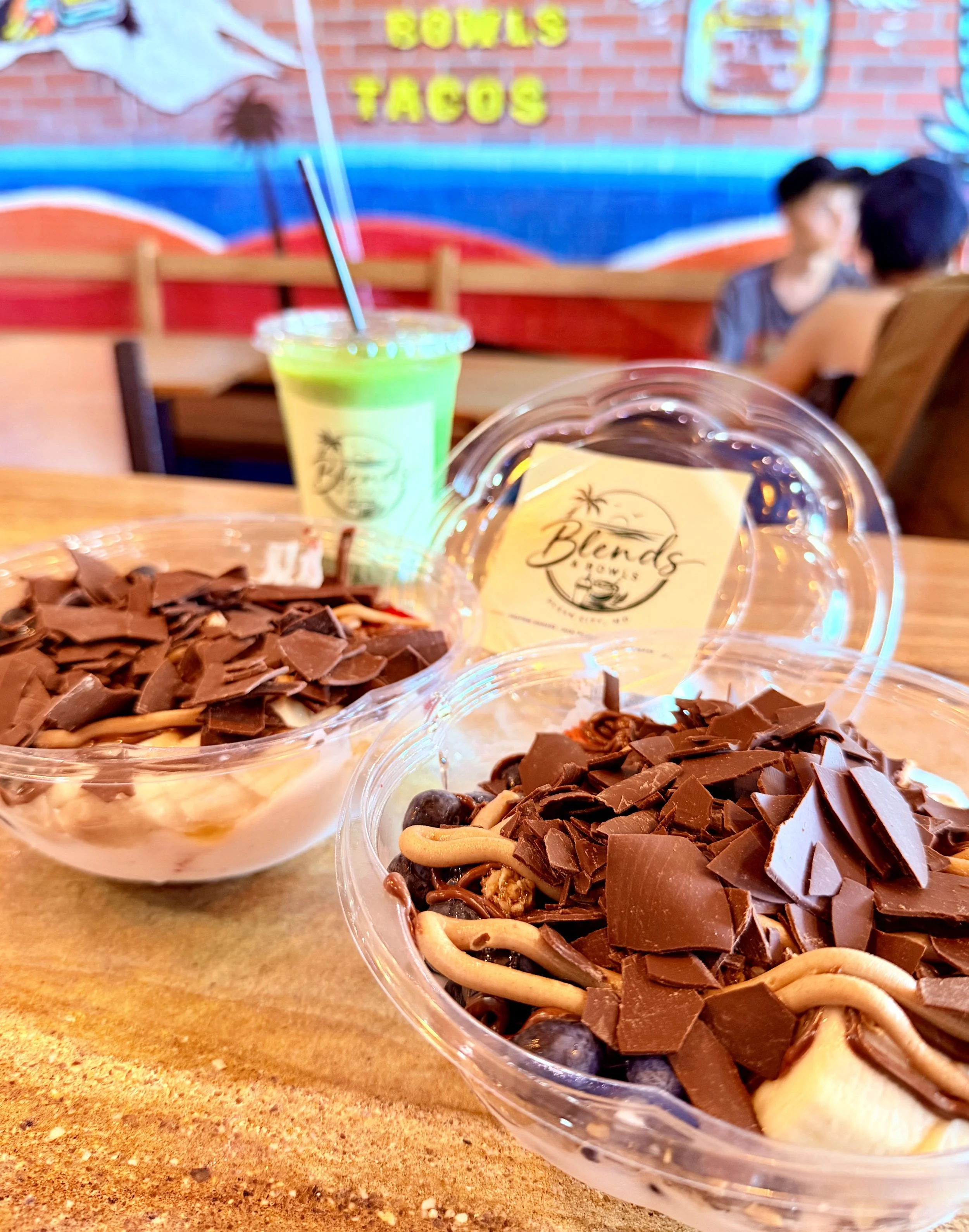 Close-up of chocolate dessert cups topped with chocolate shavings and nuts on a wooden table. In the background, a blurred scene of a restaurant with a brick wall and people seated, along with neon signs indicating 'BOWLS' and 'TACOS'.