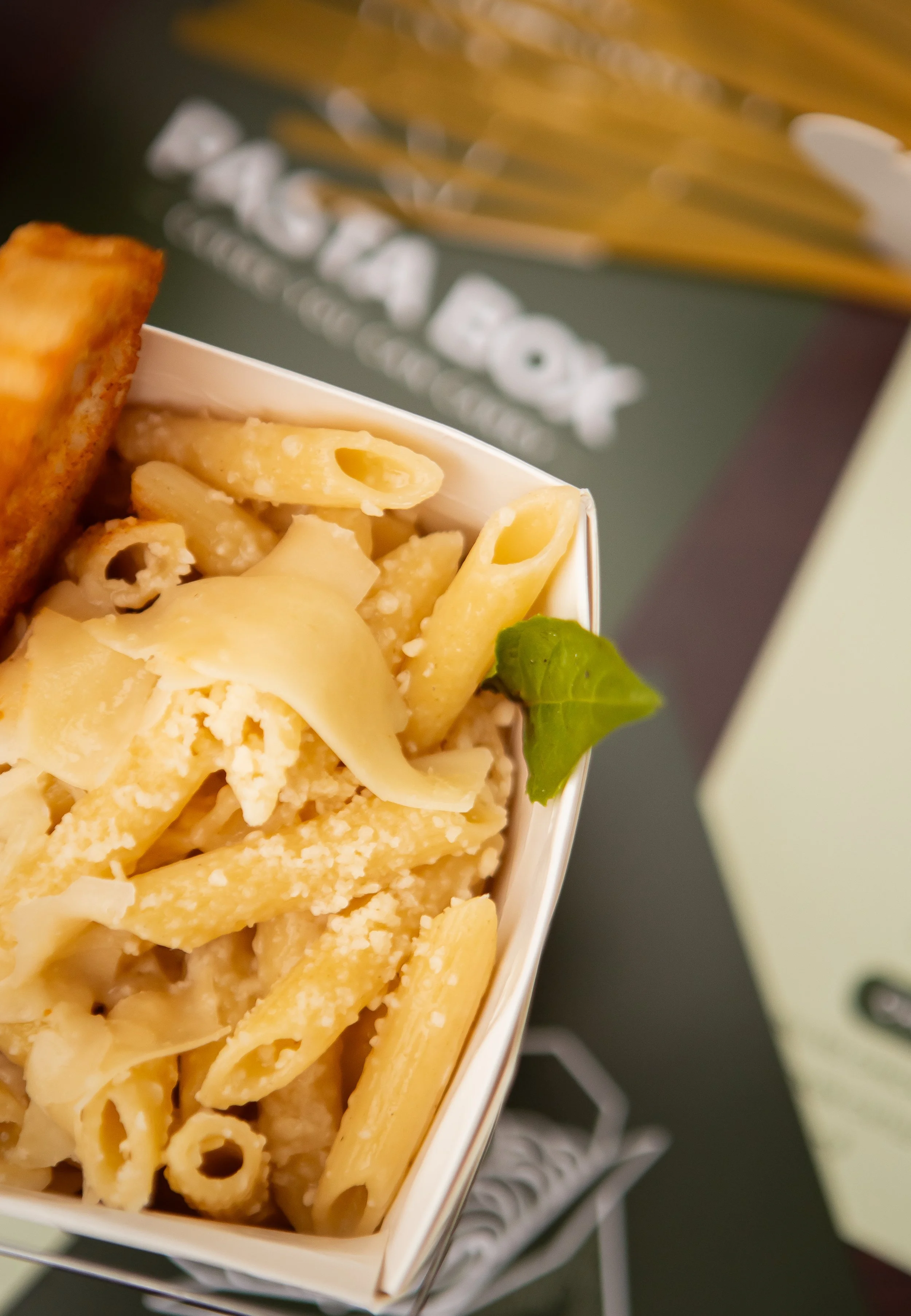 Close-up of a box of cooked pasta, including penne and other shapes, with grated cheese and a green basil leaf on top.