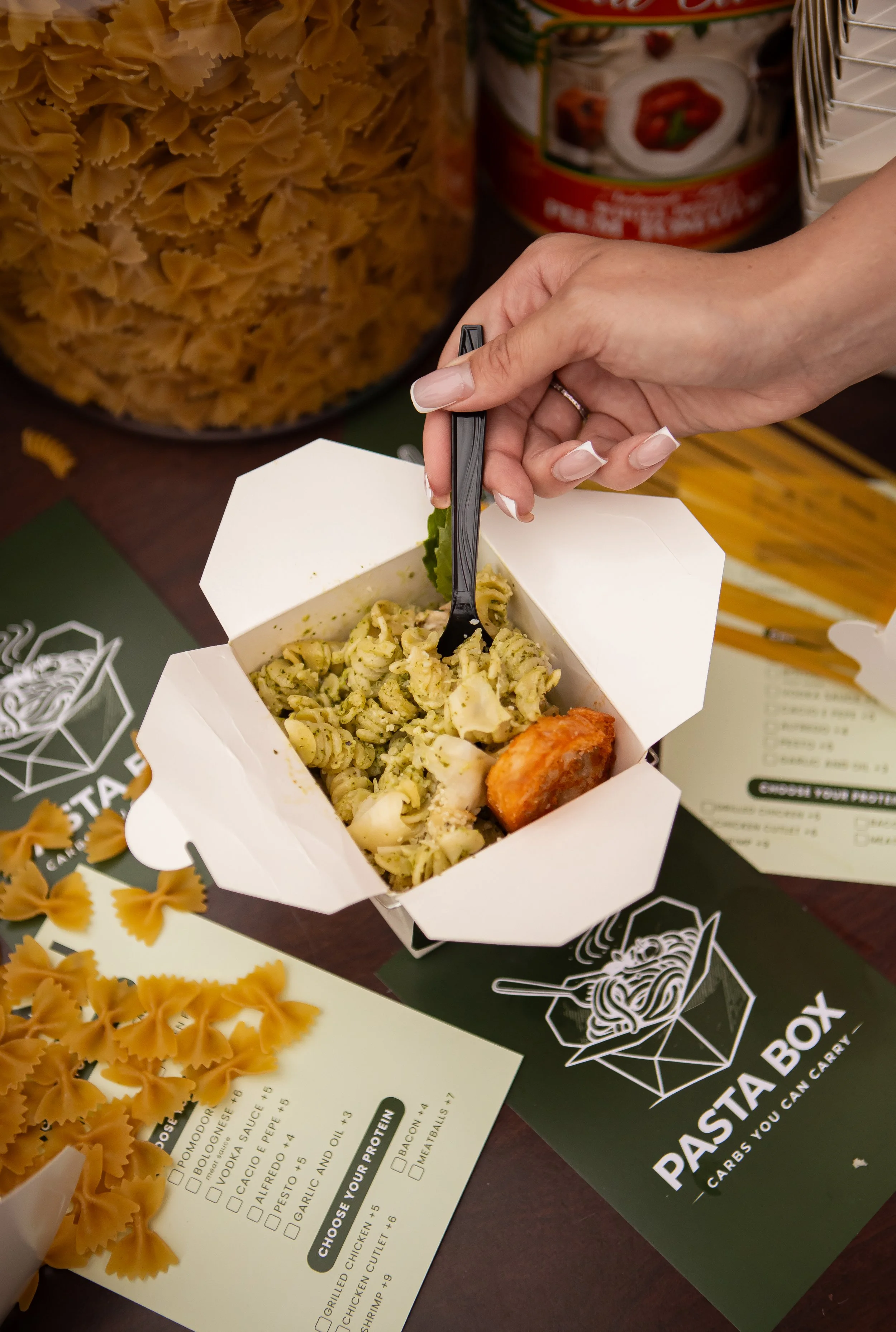 A person holding a white takeout box with pasta and a piece of fried chicken, with a black fork in the box. In the background are pasta, canned food, and a menu.