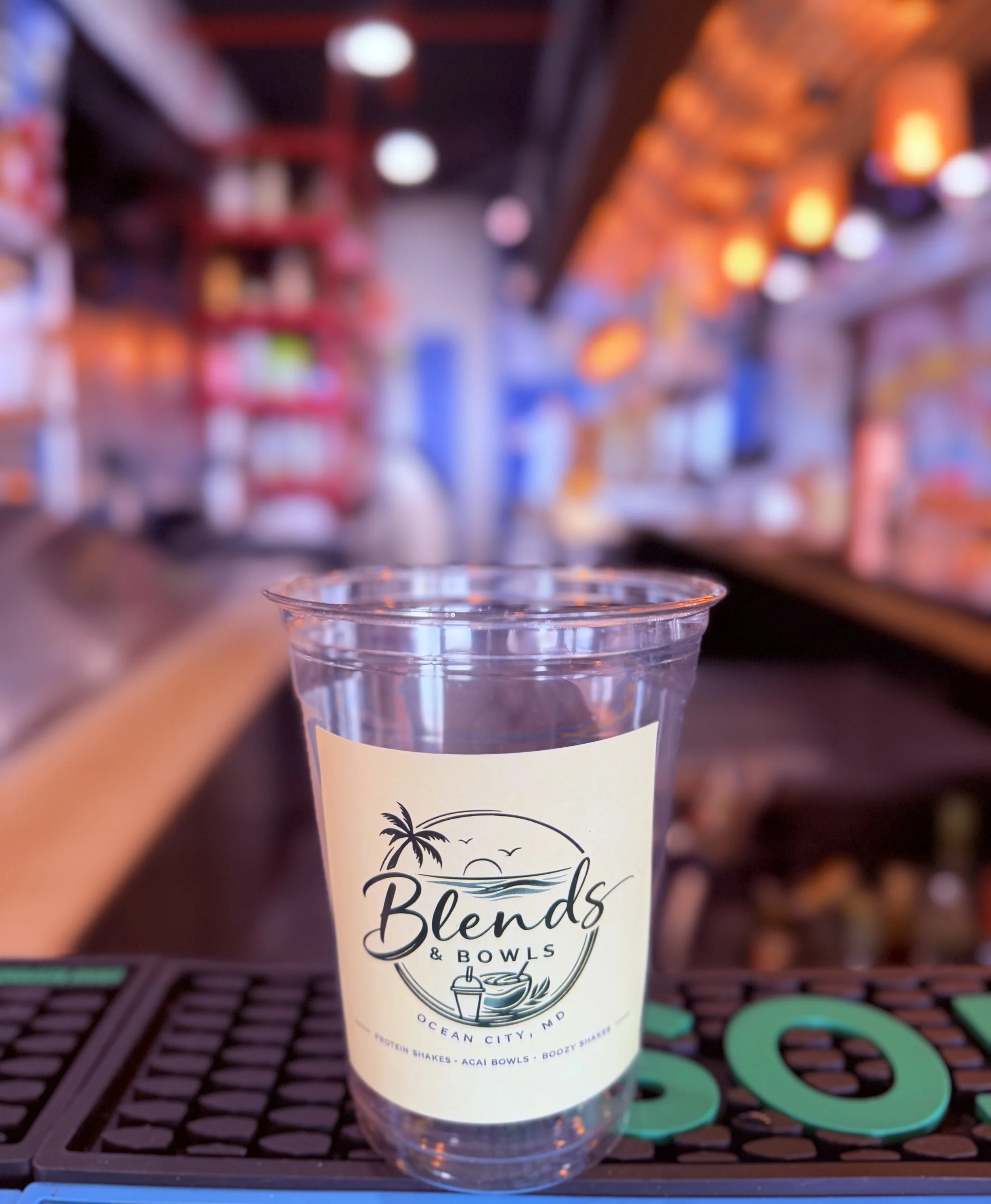 A clear plastic cup with a beach-themed label, held by a person with a finger visible in the foreground. The background is a blurred indoor setting with orange and white lights. The label on the cup says "Beach Blends & Bowls" with a graphic of a pal