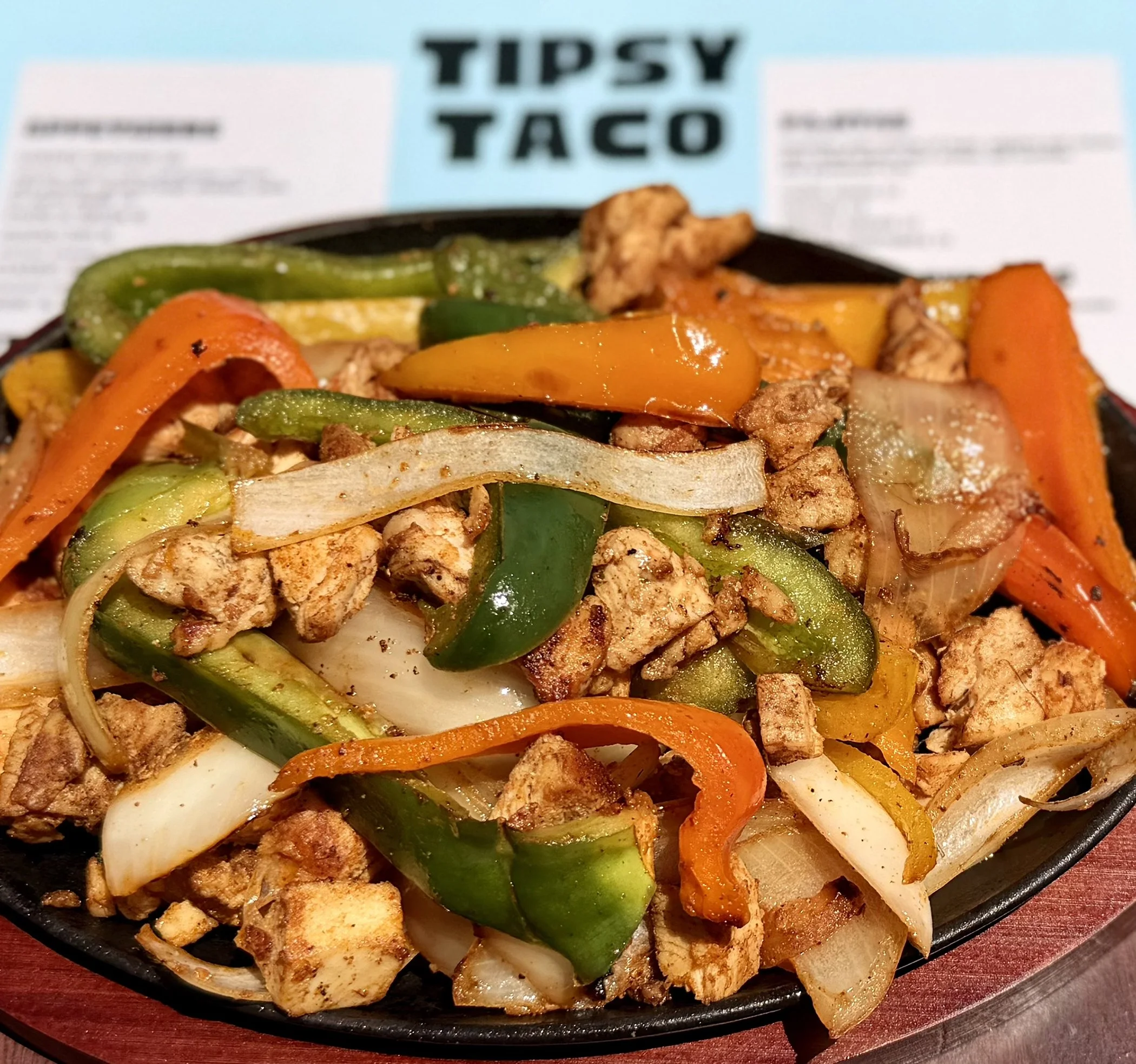 A sizzling cast iron skillet filled with grilled vegetables and pieces of seasoned chicken, with a menu for Tipsy Taco in the background.
