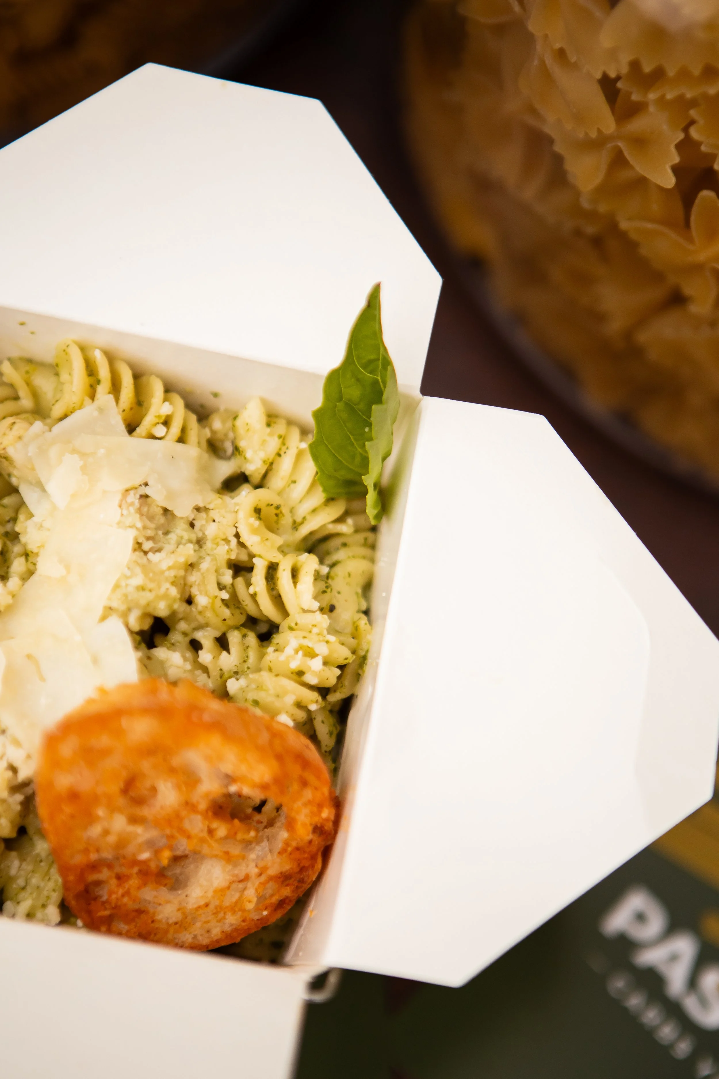 Close-up of a takeaway food container filled with pasta salad, topped with cheese and a leaf of basil, and a fried food item, possibly a croquette or nugget.