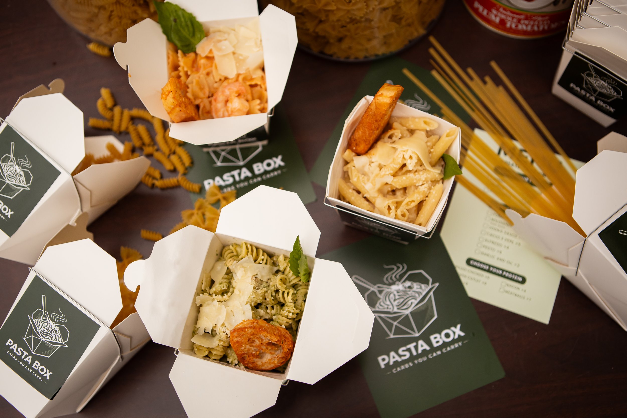 Several white takeout boxes filled with different types of pasta, some topped with cheese and one with a piece of breaded chicken or seafood, placed on green paper with a logo, on a dark table with scattered fusilli pasta and some canned foods in the background.