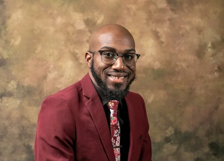 A smiling man with glasses, a bald head, and a beard, wearing a maroon suit and floral tie, against a brown mottled background.