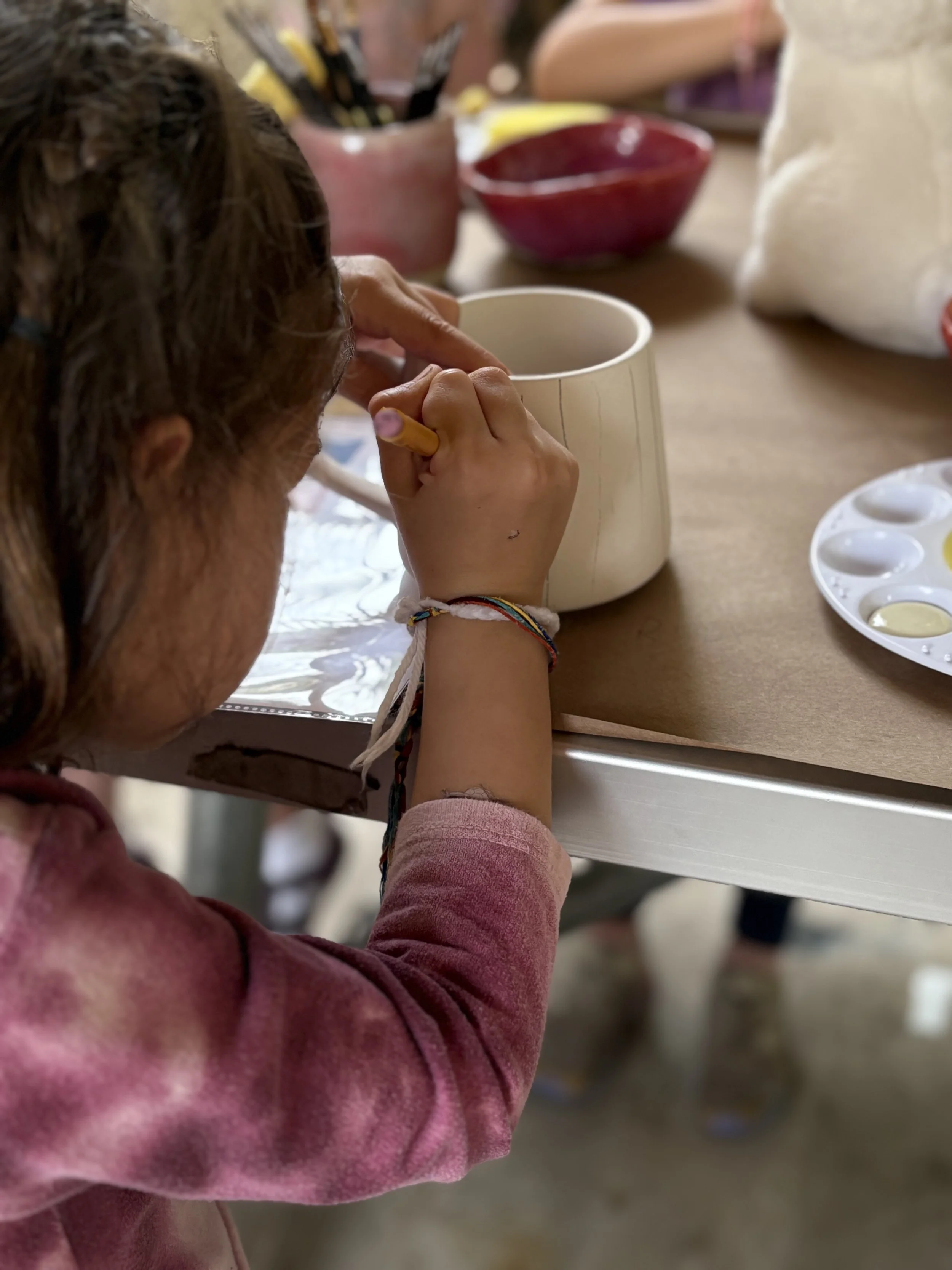 Child painting on a mug with yellow paintbrush, sitting at a table with bowls of paint and art supplies.
