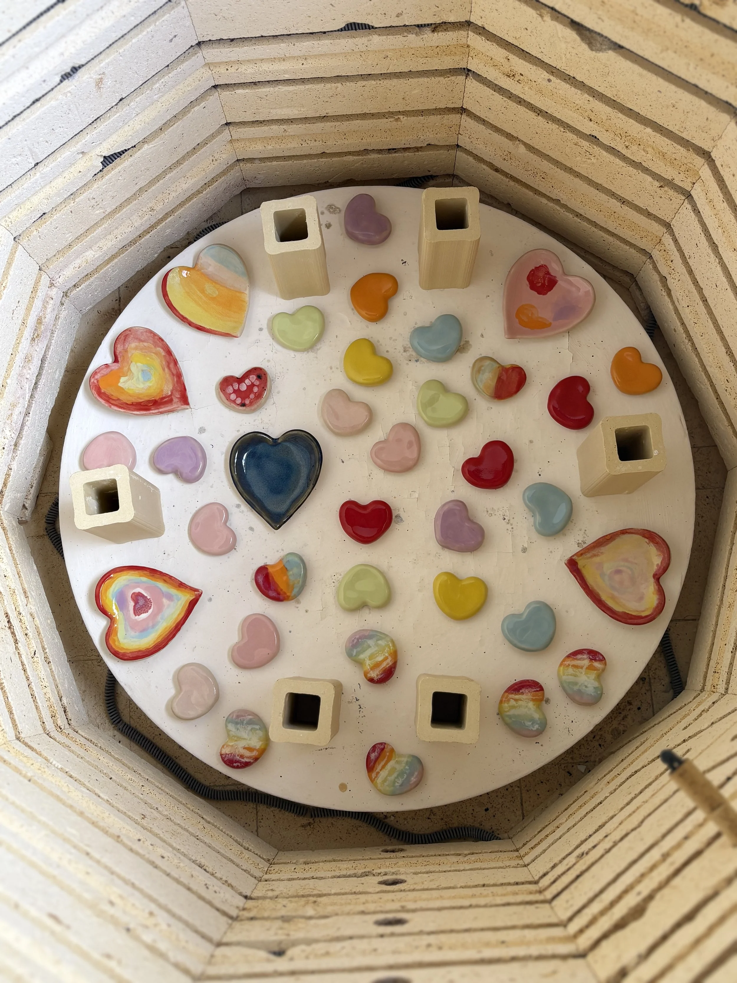 A round display of colorful heart-shaped and abstract glass art pieces within a wooden hexagonal frame.