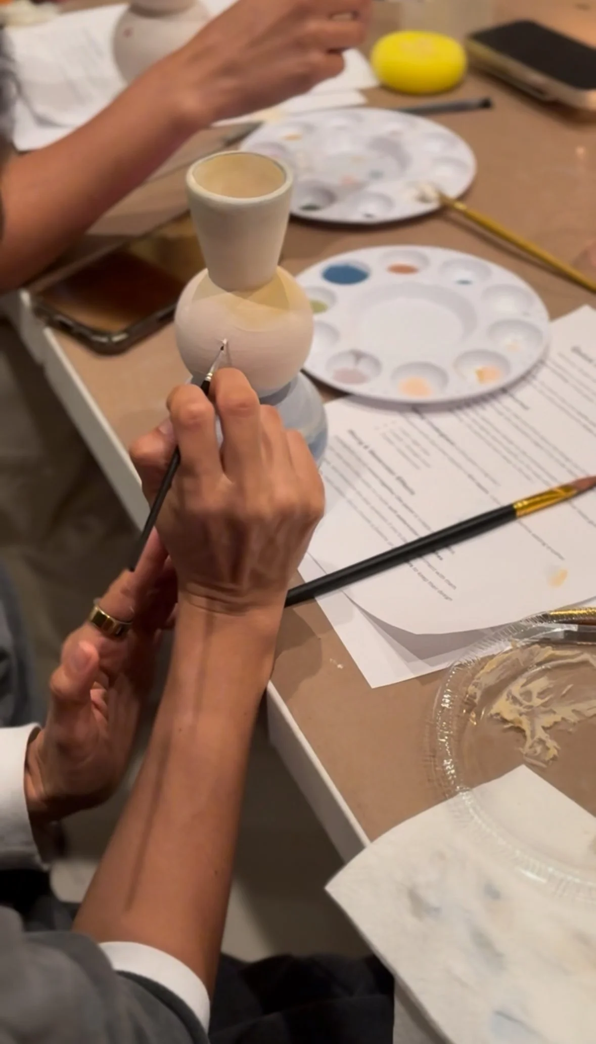 Close-up of a person painting a small ceramic vase, with painting supplies and a plate of mixed watercolor paints on the table.