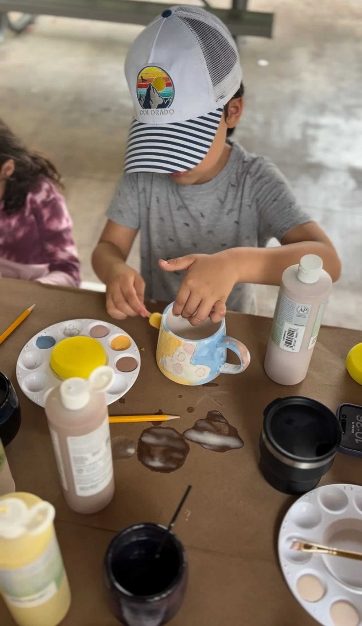 A boy wearing a Colorado hat decorates a mug with paint while sitting at a table with art supplies and paint bottles.