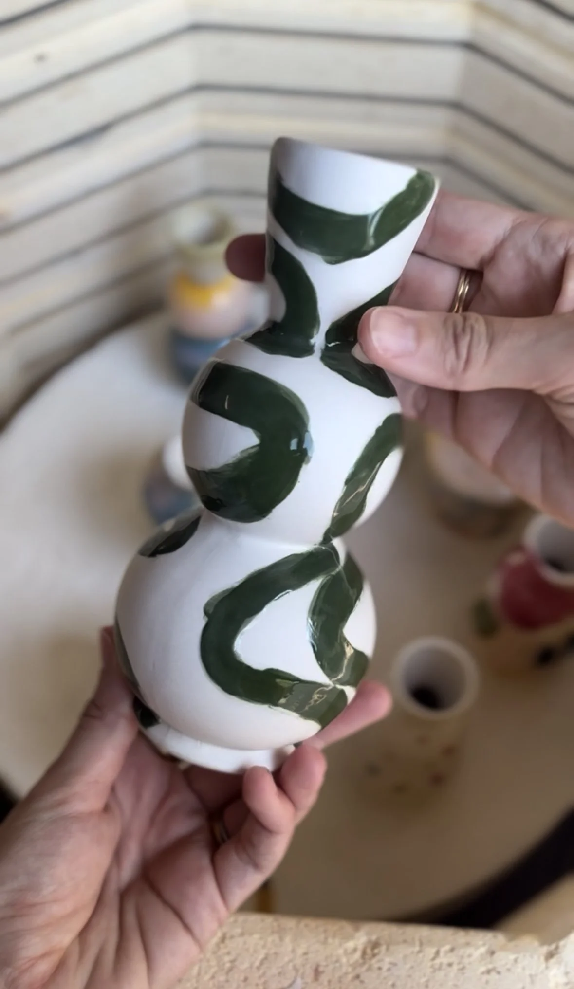A person holding a white ceramic vase with green abstract brushstrokes. Other similar vases are in the background on a round table.