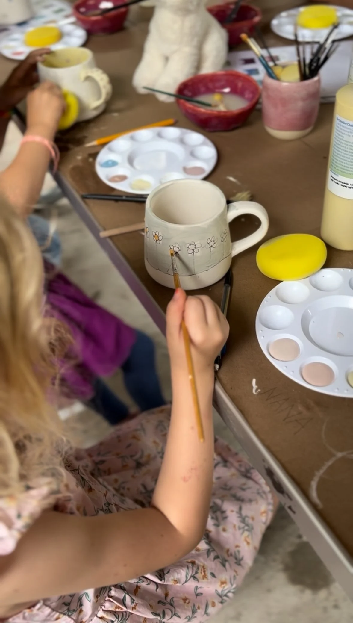 A child painting a flower design on a ceramic mug, surrounded by painting supplies, including paint palettes, brushes, and painted mugs, in an art studio setting.