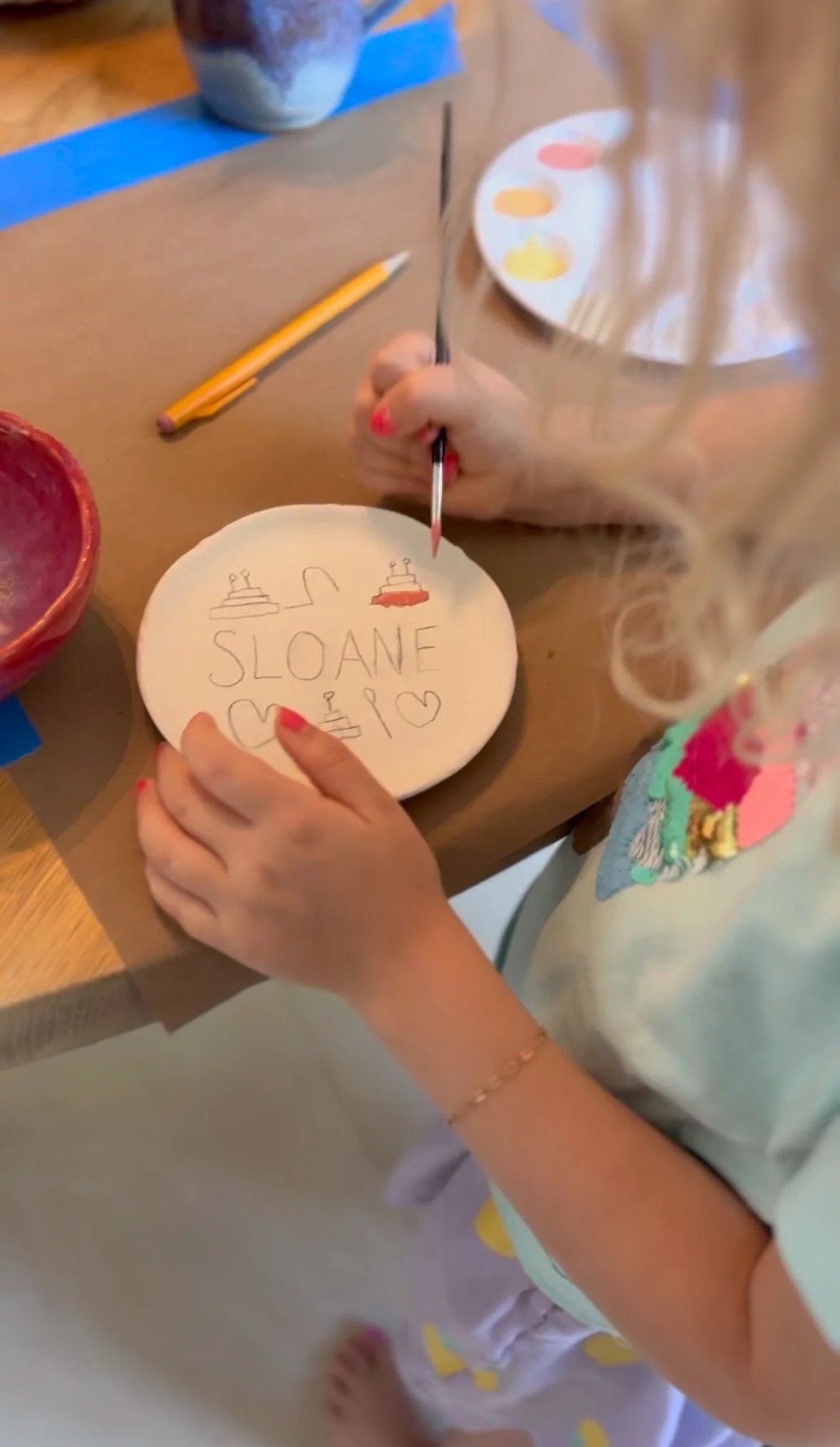 A young girl with blonde curly hair, painting a decorated oval ceramic object with the name 'SLOANE' and hearts using paintbrushes. Art supplies and a palette are visible on the table.