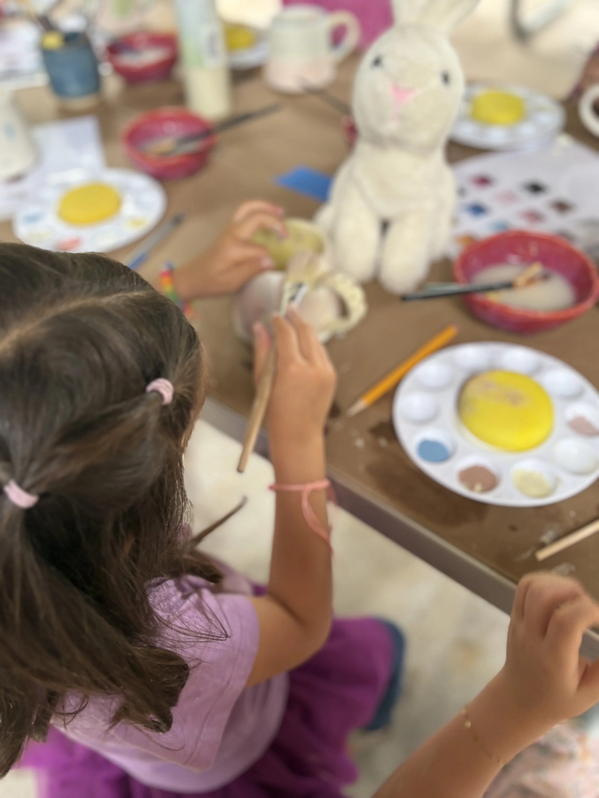 A young girl painting a white plush bunny toy at a craft table with various paints and brushes.