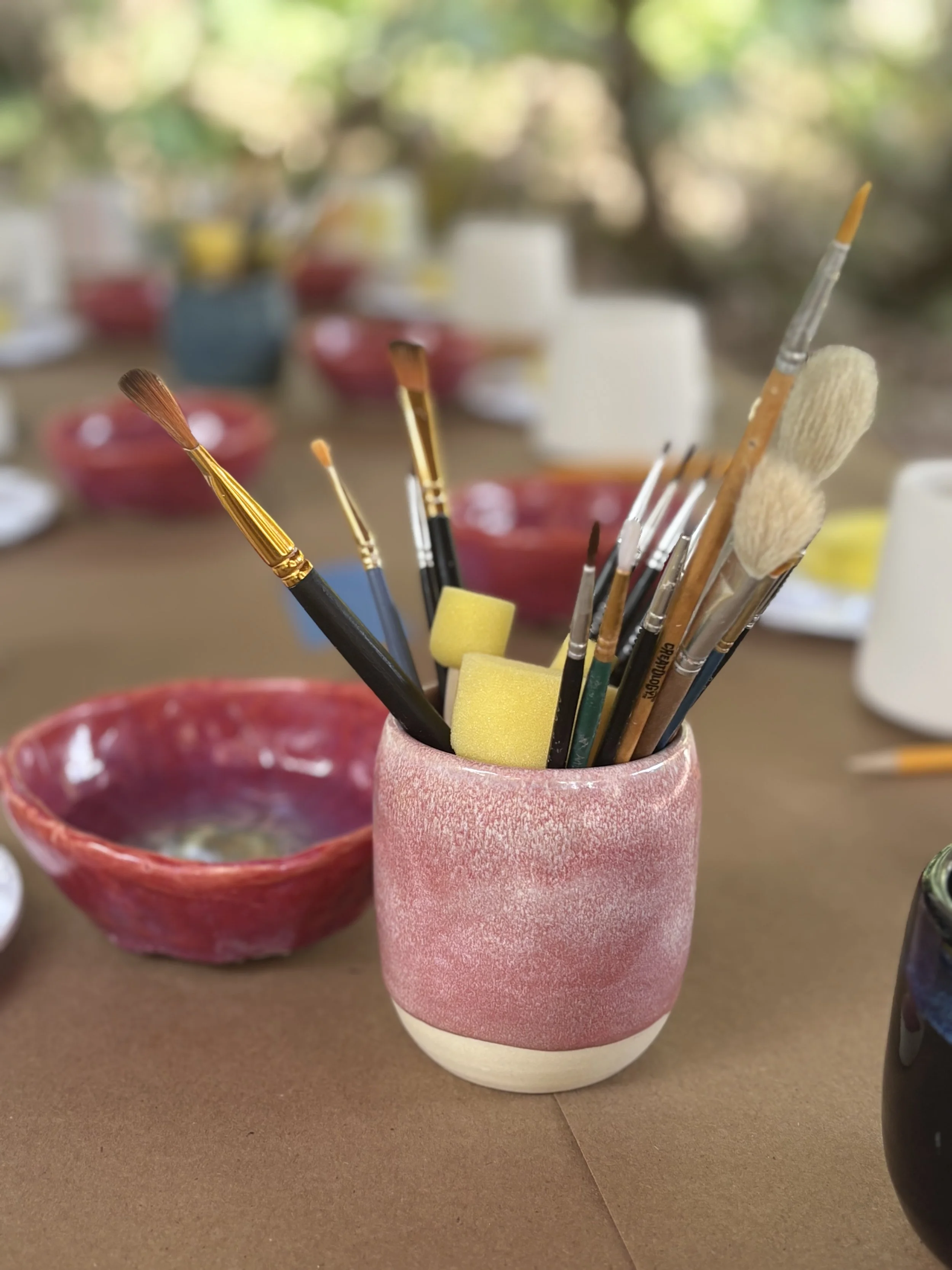 A pink ceramic cup holding various paintbrushes and sponges on a table with other bowls and objects in the background.