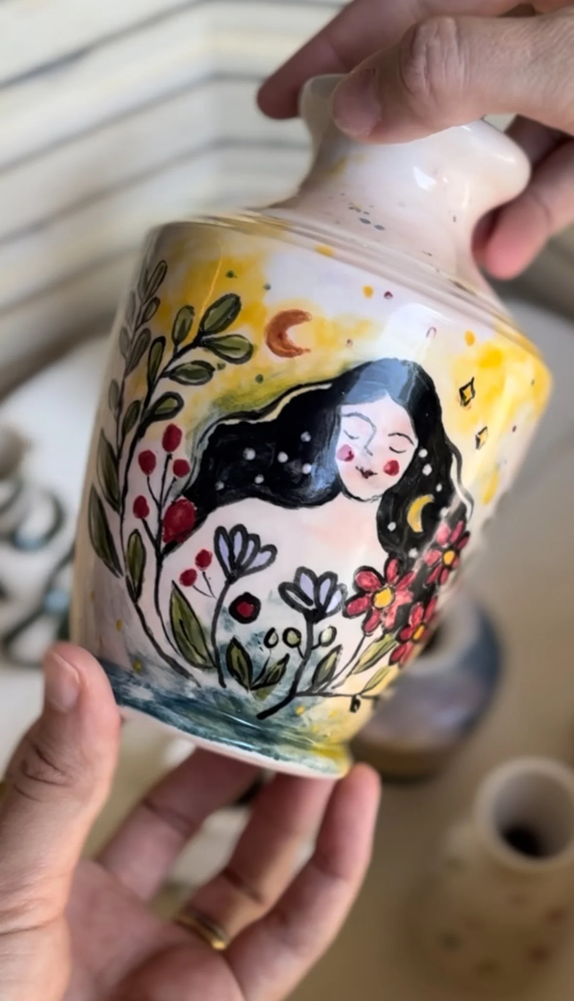 A person holding a ceramic jar with a colorful, artistic design featuring a woman's face with long dark hair, surrounded by flowers, leaves, and a background of yellow, orange, and blue hues.