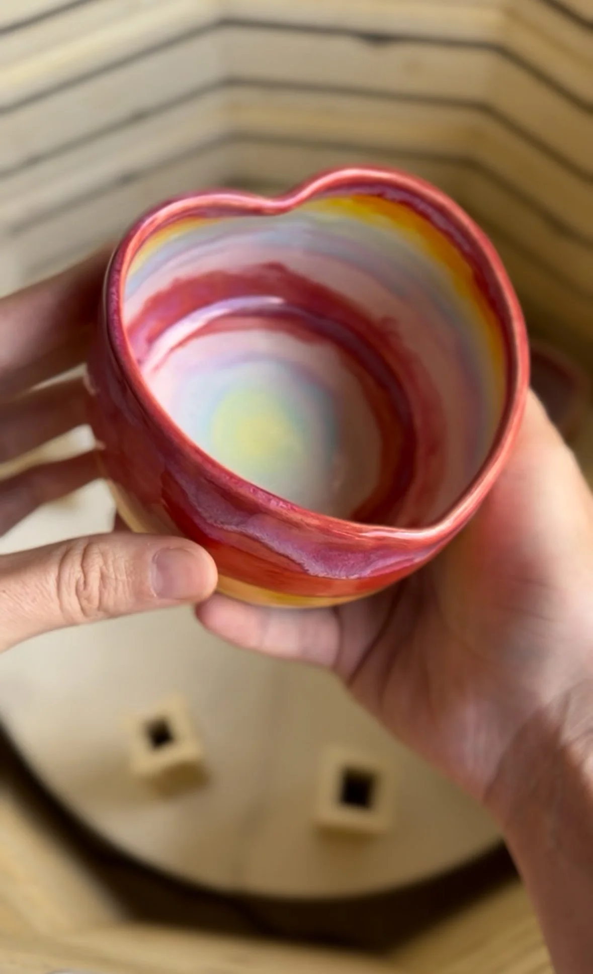 A person holding a small heart-shaped ceramic cup with colorful, swirl patterns inside, against a background with a wooden surface and small square objects.
