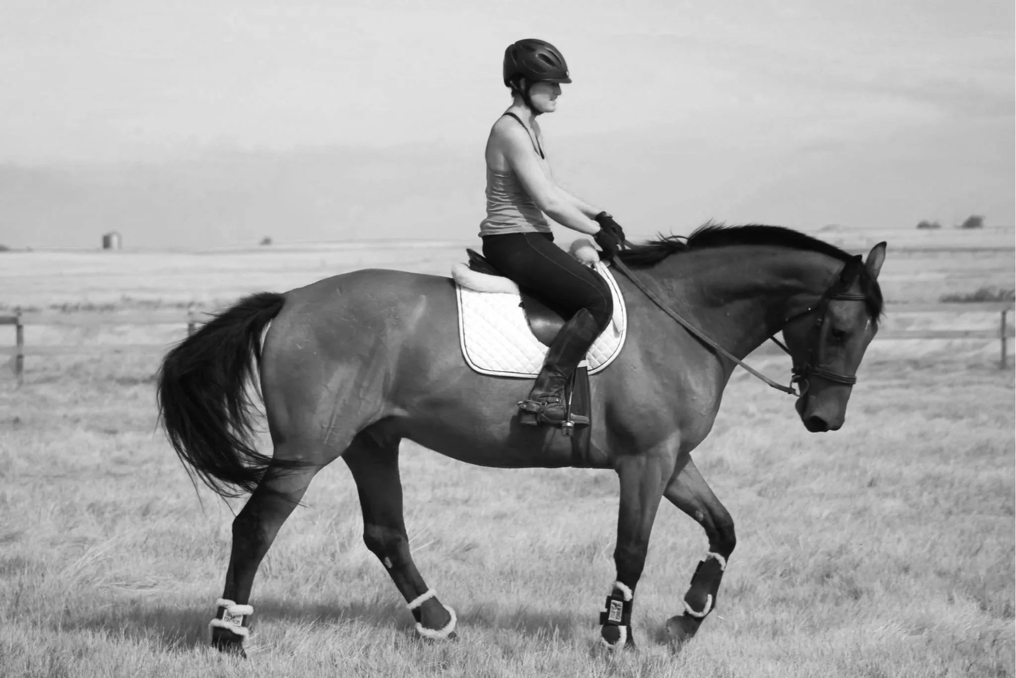 A woman riding a horse in an open field, wearing a helmet, tank top, and riding gear, in black and white.