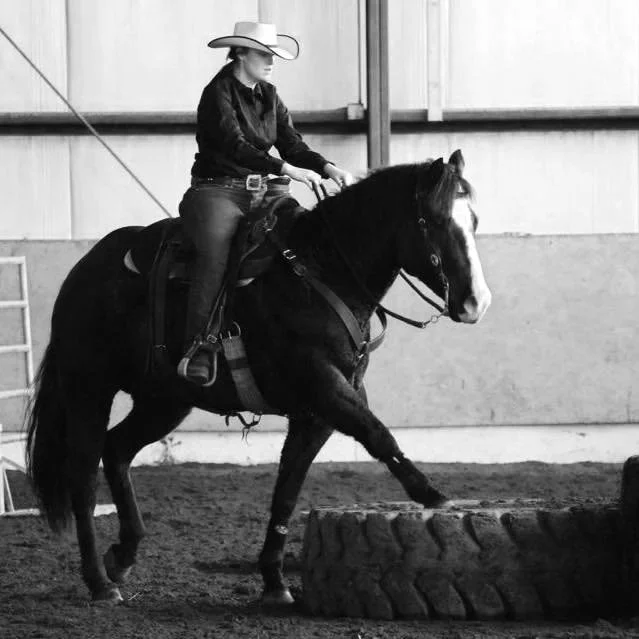 A person wearing a cowboy hat riding a horse inside an indoor arena, approaching a large tire obstacle.