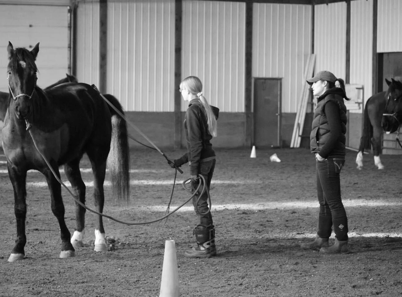 Two women practicing horseback riding in a spacious indoor riding arena. One woman holds a horse, the other observes.
