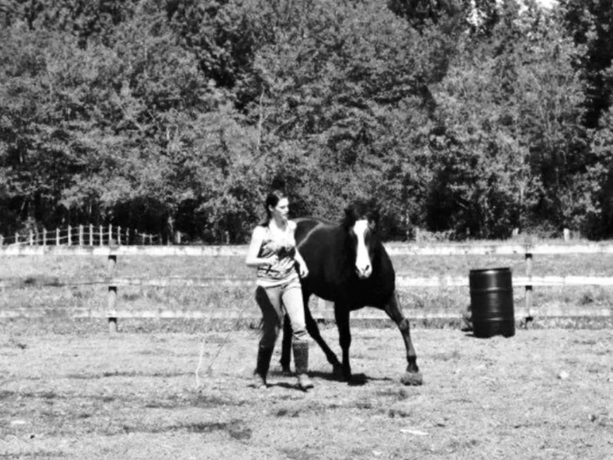 A woman leading a horse in an open field with trees in the background, black and white