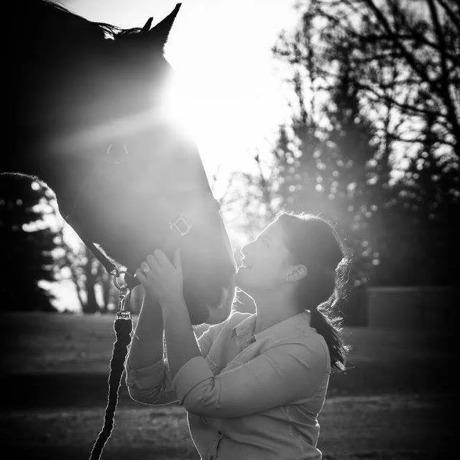 A woman is holding a horse's face close to hers, touching noses, outdoors with trees in the background, backlit by the sun, black and white photo.