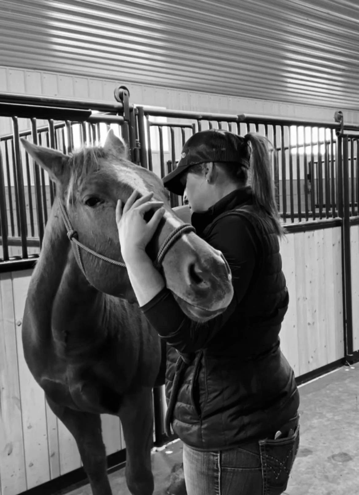 A woman petting a horse in an indoor stable. The woman is wearing a baseball cap and a jacket, and the horse is standing close to her.