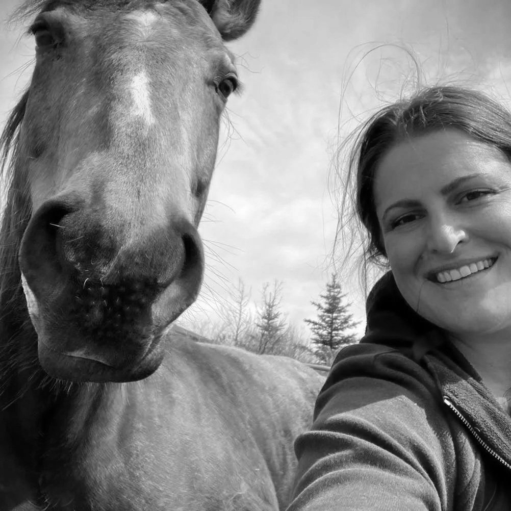 A woman taking a selfie with a large horse outdoors, with trees and cloudy sky in the background, in black and white.