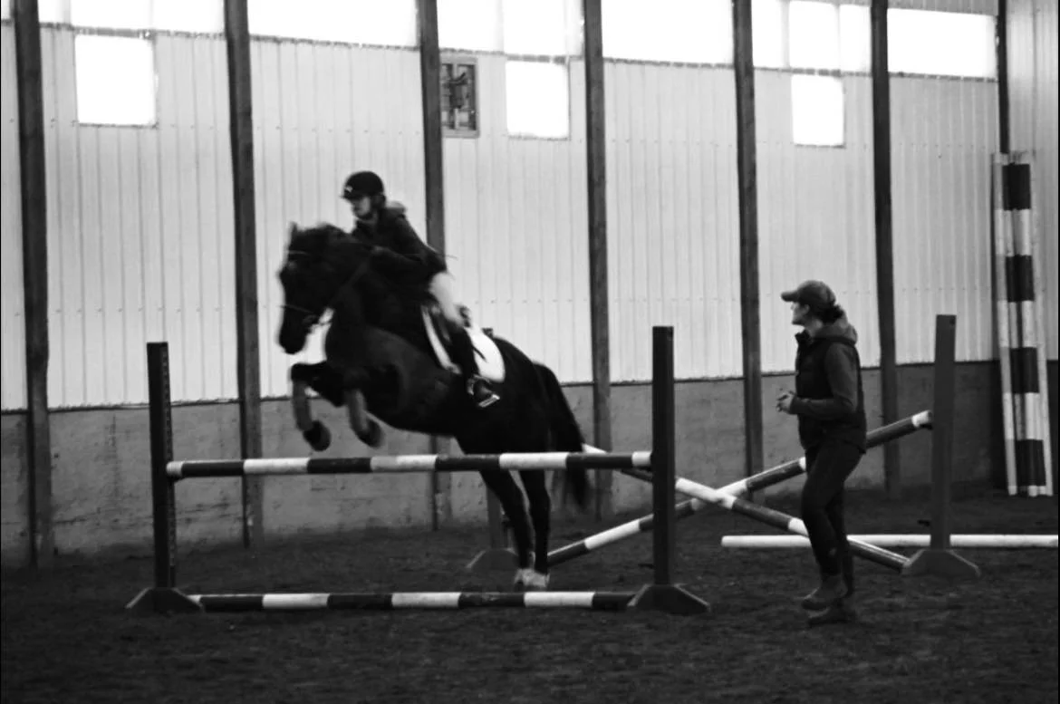 A person riding a horse jumping over an obstacle while a trainer guides them in an indoor arena.