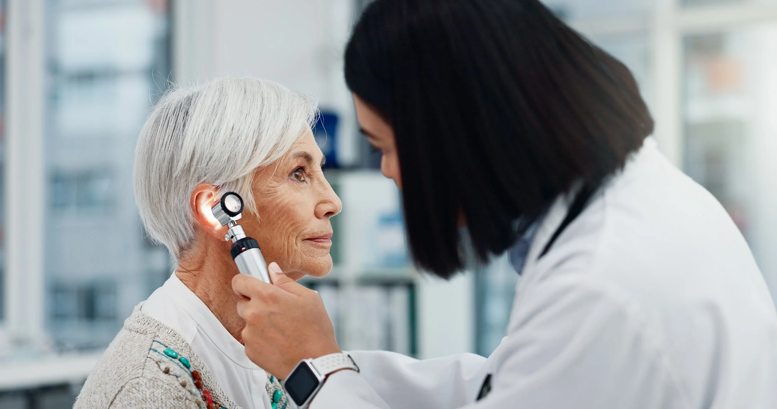 Doctor examining an elderly woman's ear with otoscope in a medical office.