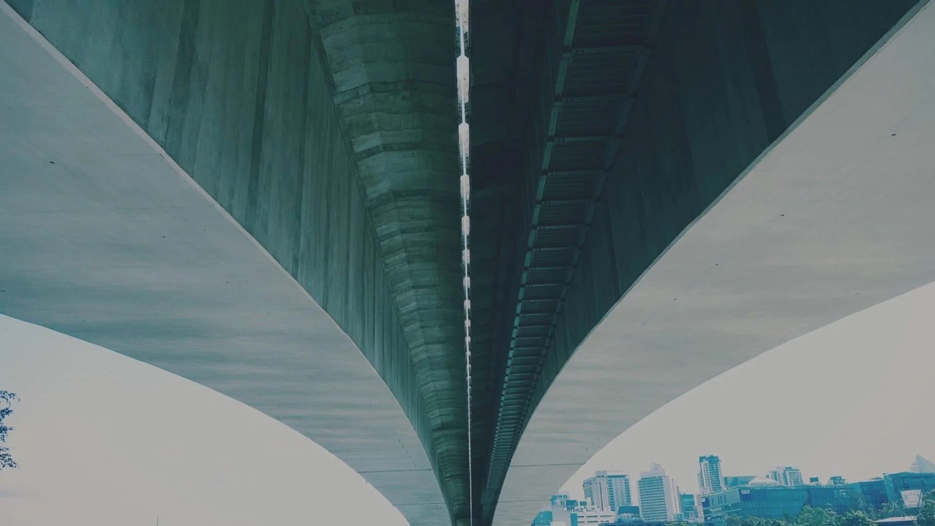 Vista de abajo de un puente elevado de concreto con estructura metálica en el centro y ciudad moderna en el fondo.