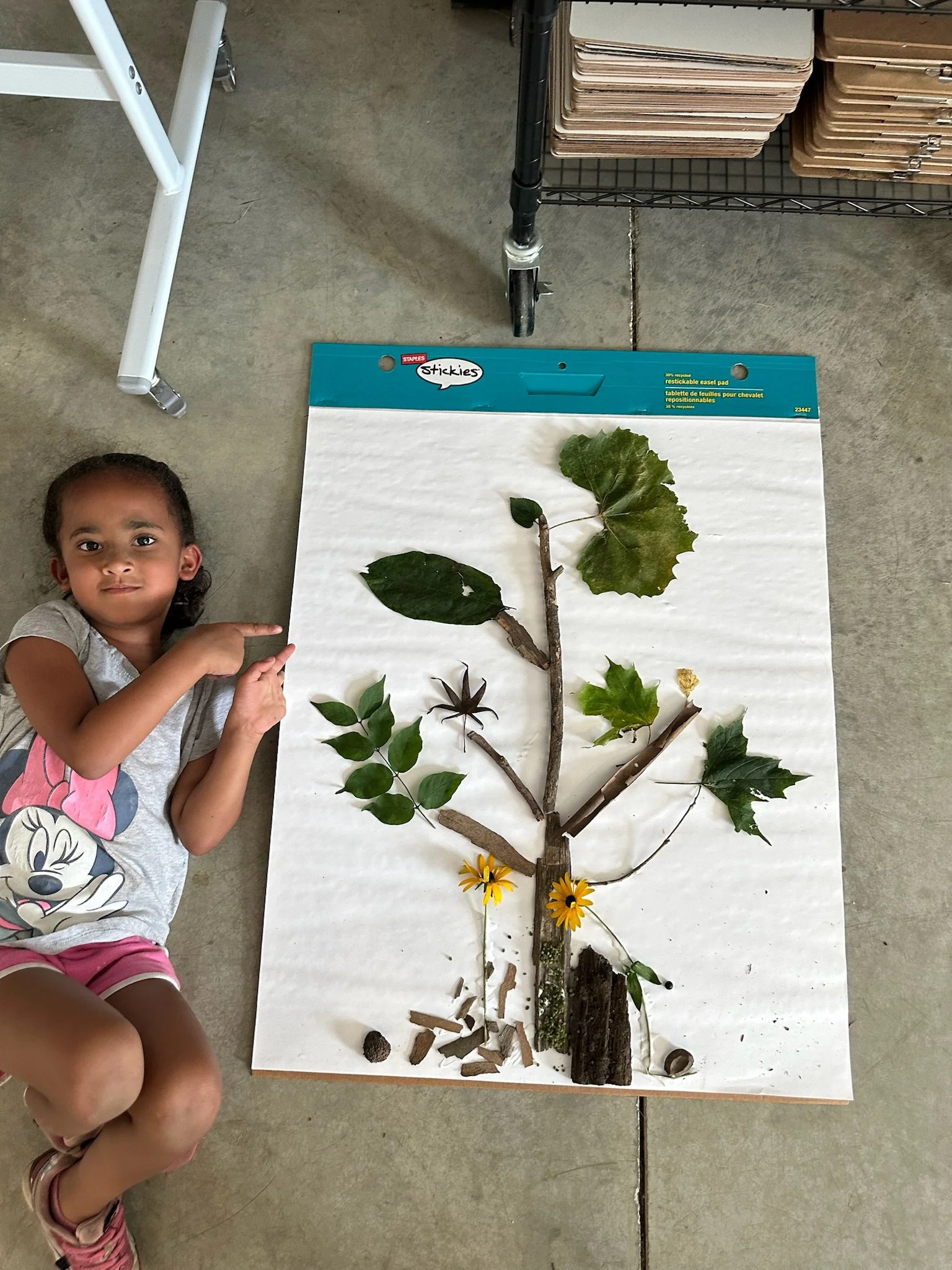 A young student smiles beside a tree-shaped nature collage made with sticks, leaves, flowers, bark, and other found materials.