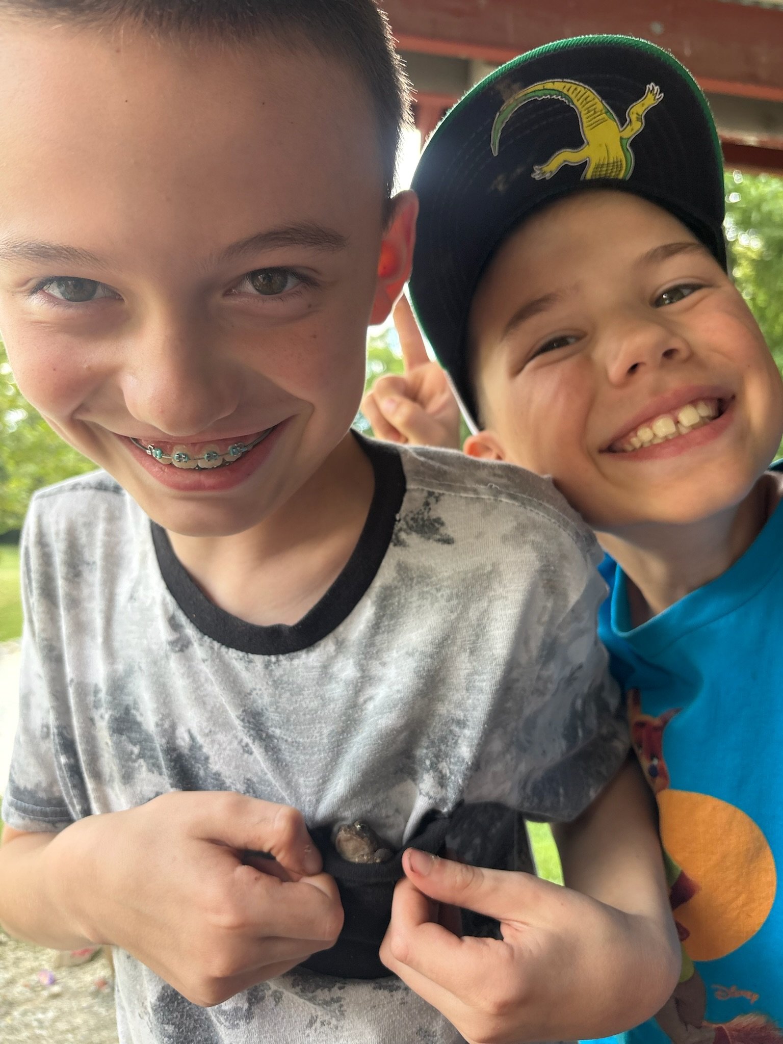 Two smiling students excitedly show a tiny frog they discovered during an outdoor nature activity.