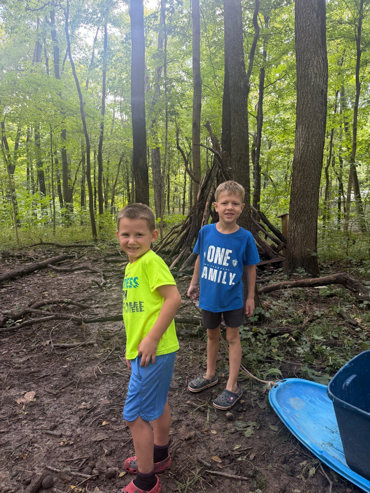 Two students stand in the woods beside a shelter they built from fallen branches leaned against a tree.