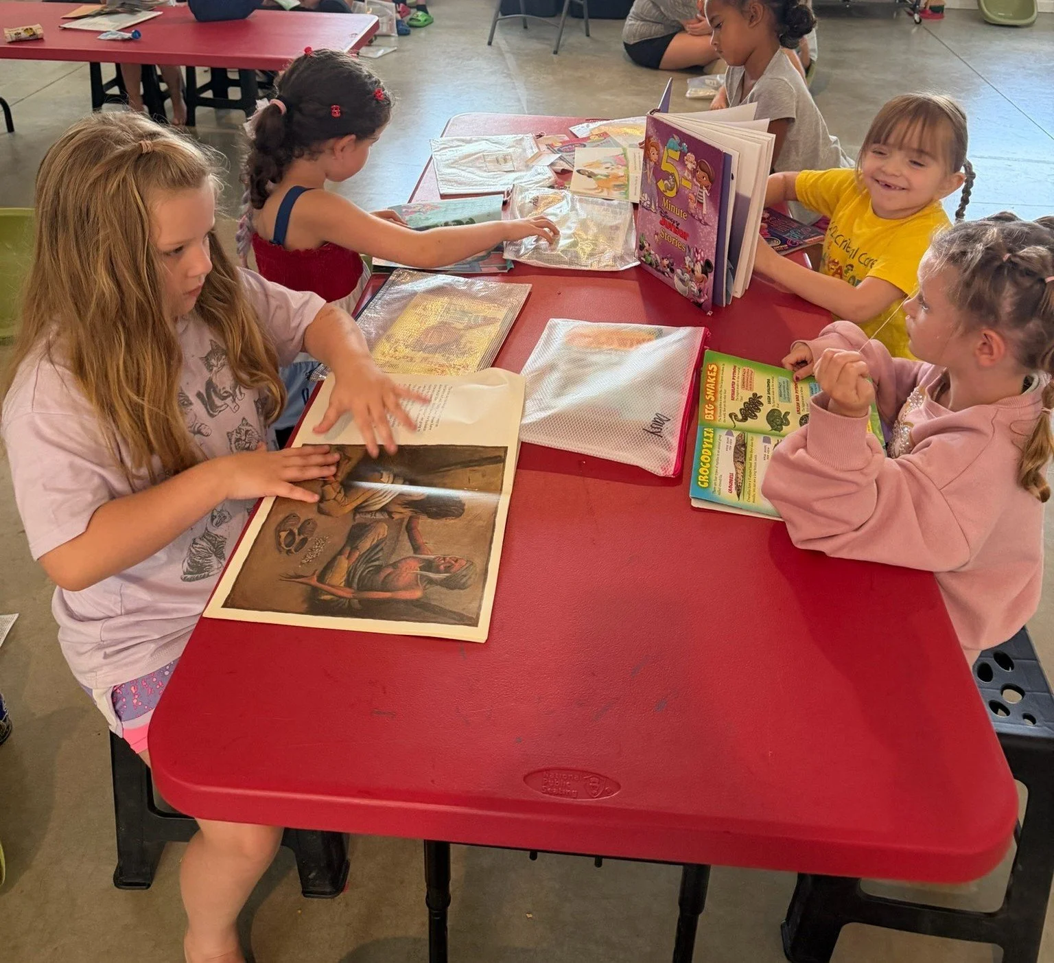 Students sit together around a table looking through books and sharing stories during an indoor activity.