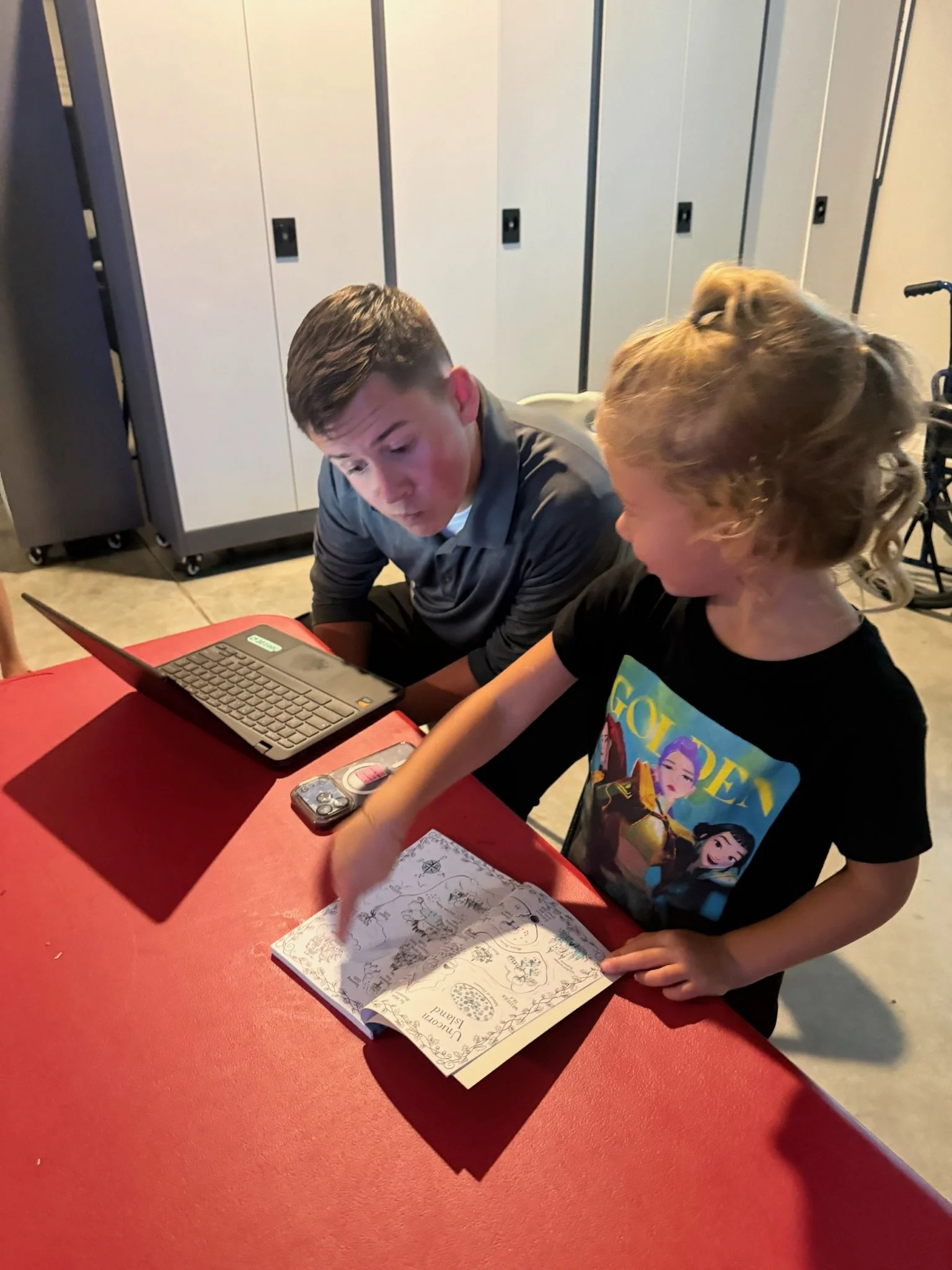 A young student points to a page in a book while an older student looks on during an indoor learning activity.