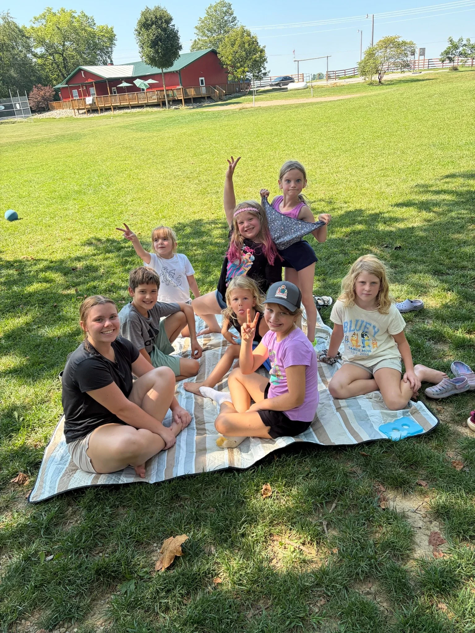 Students sitting on a blanket outside in the grass. A big red building behind them. They are smiling and looking at the camera.