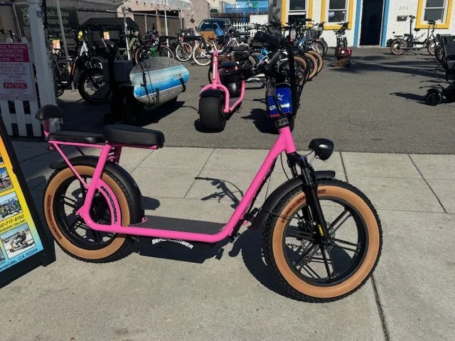 Pink electric bicycle with brown tires parked on sidewalk at a bike rental station.