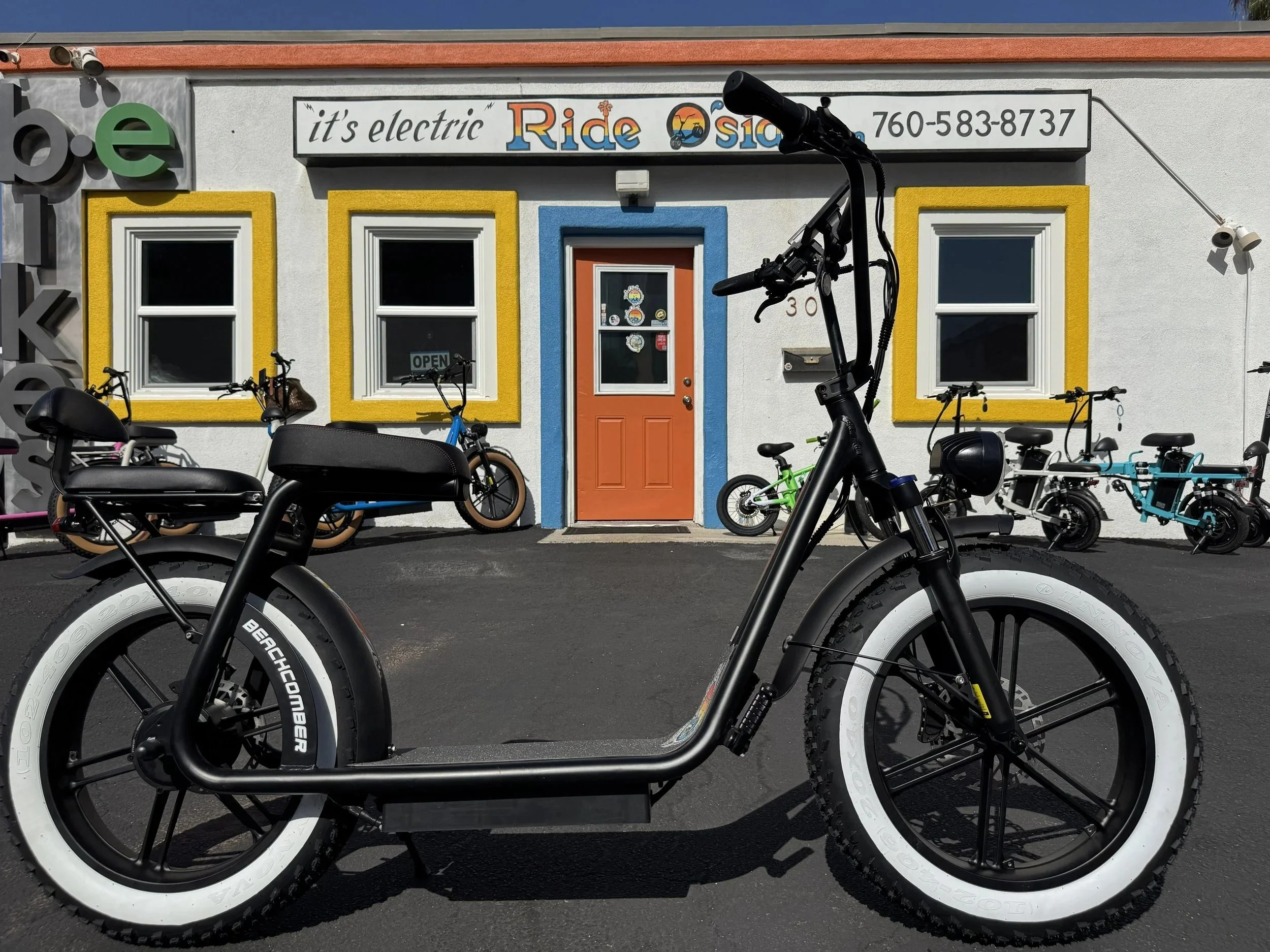 Black electric bicycle with white tires parked in front of a colorful storefront with yellow, blue, and orange accents. The storefront has a sign that reads 'it's electric Ride Oasis' and a phone number. Other bicycles are also parked in front of the store.