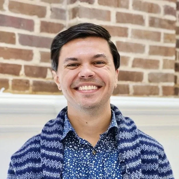 Connecting Heals clinical director & founder, Steven Gilipin, wearing a blue patterned shirt and a knit sweater, standing in front of a brick wall.