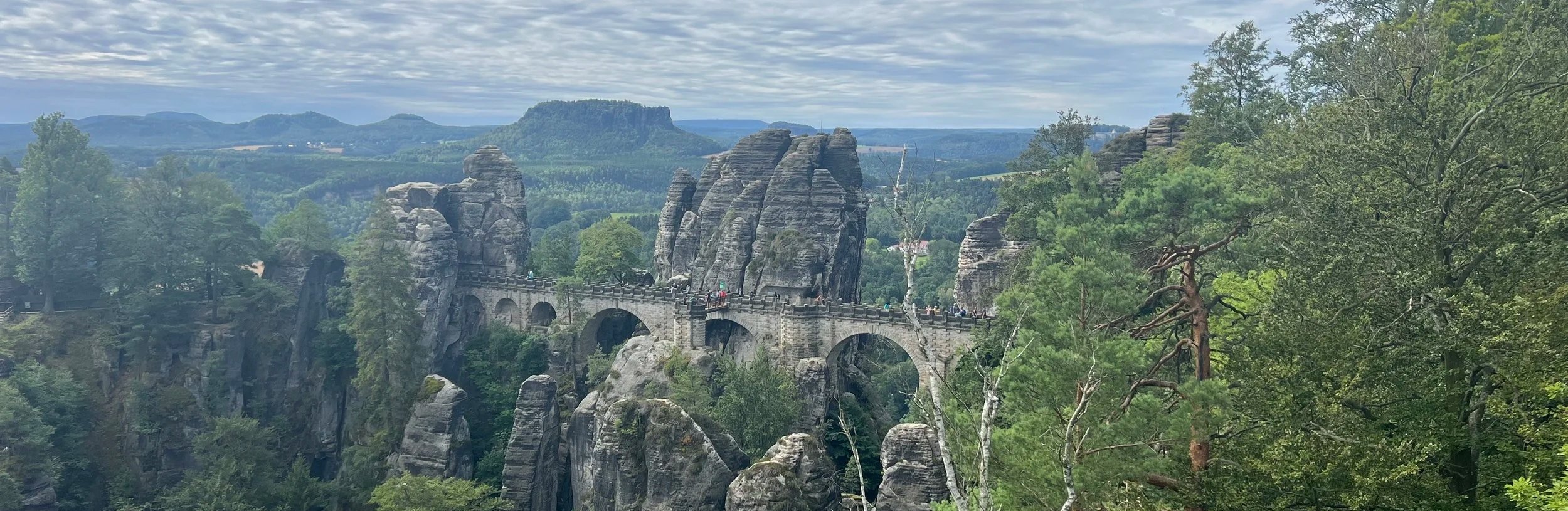 A peaceful stone bridge over rugged rocks with people walking slowly. Green trees and rolling hills stretch into a soft cloudy sky. Nature’s calm contrasts with the bridge’s strength.