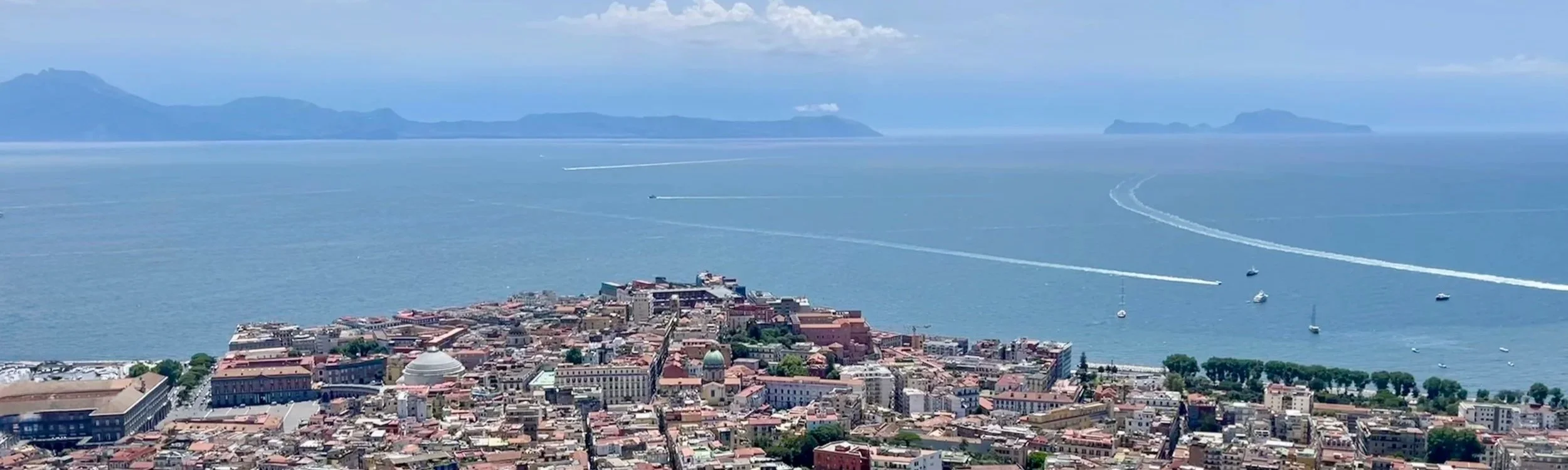 Scenic point extending into the bay of Naples with colorful buildings, boats, and mountains under soft clouds.